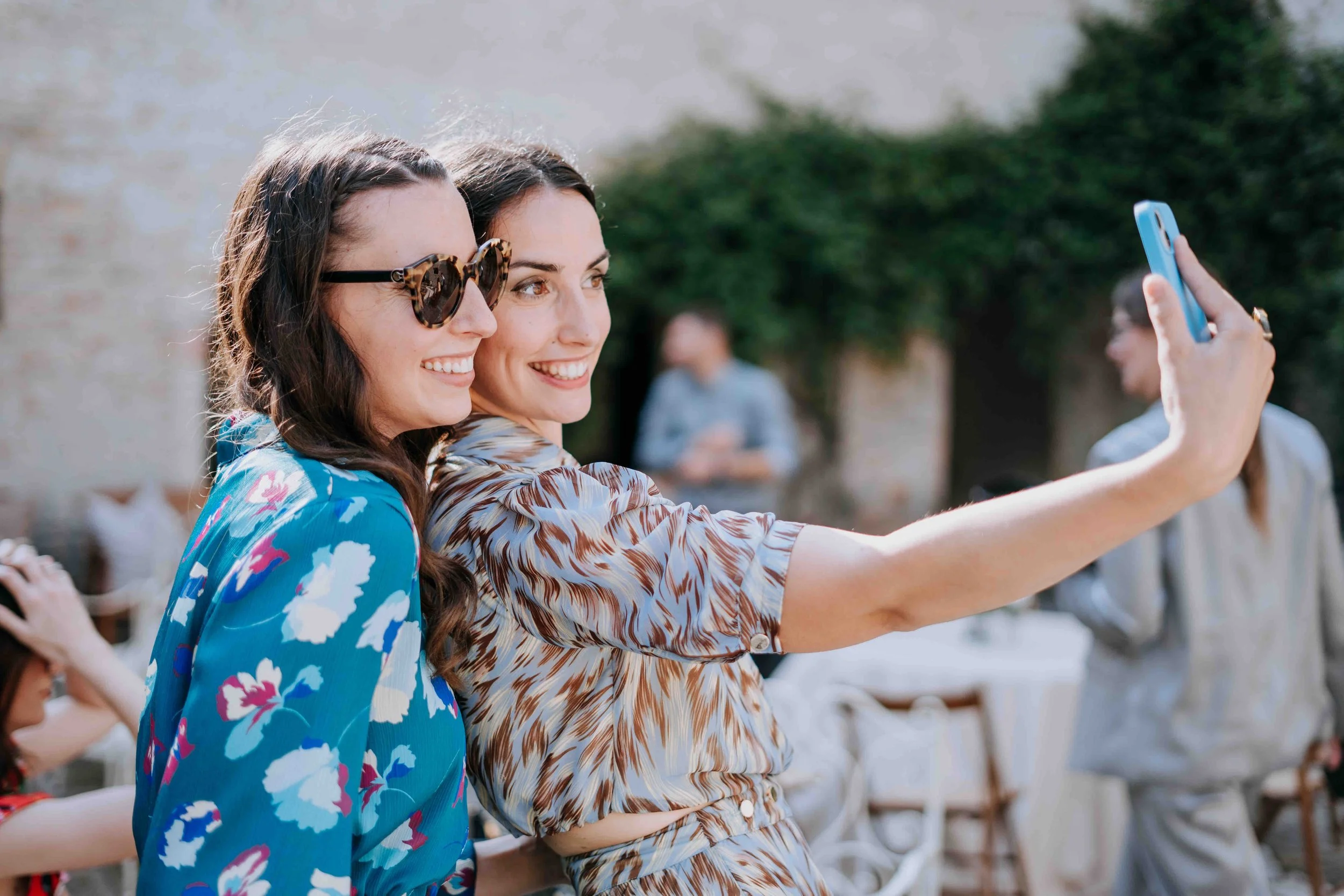 Two women smiling and taking a selfie together outdoors at a social gathering, with other people in the background.