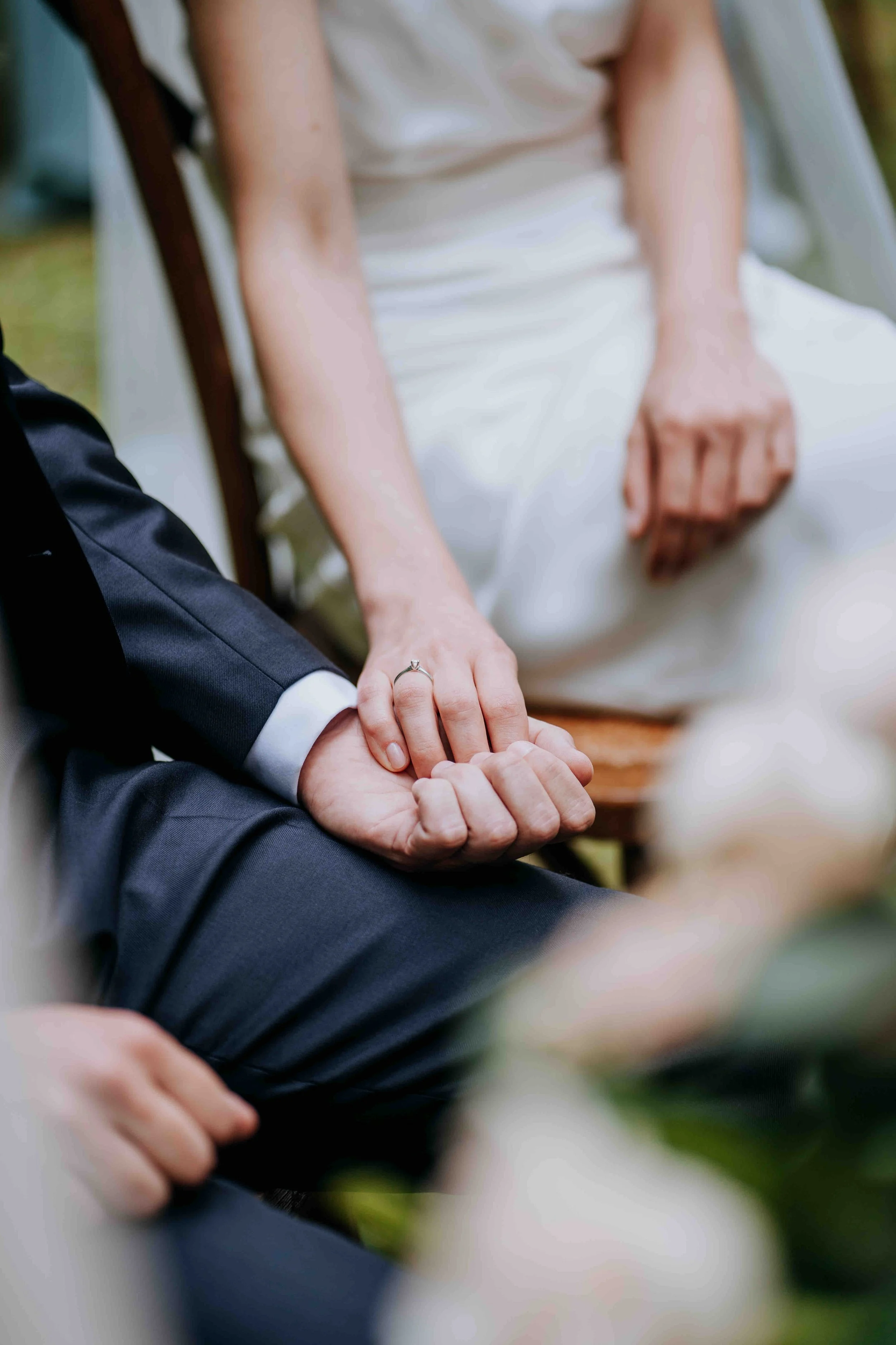 Close-up of a couple holding hands during a wedding ceremony, with the woman's wedding ring visible.