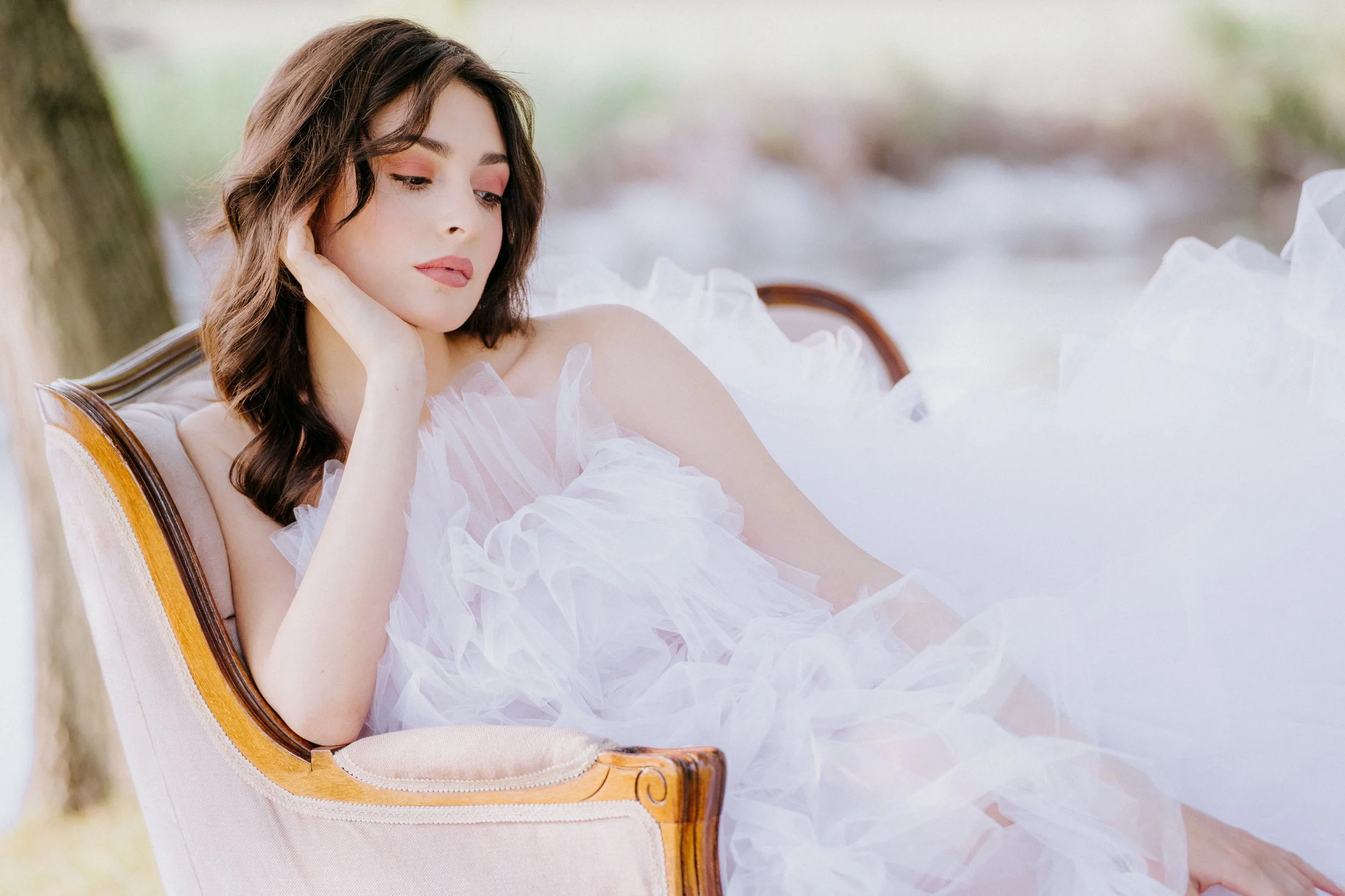 A woman with wavy brown hair, wearing a white, ruffled dress, resting her head on her hand while sitting on a vintage-style sofa outdoors near a tree.