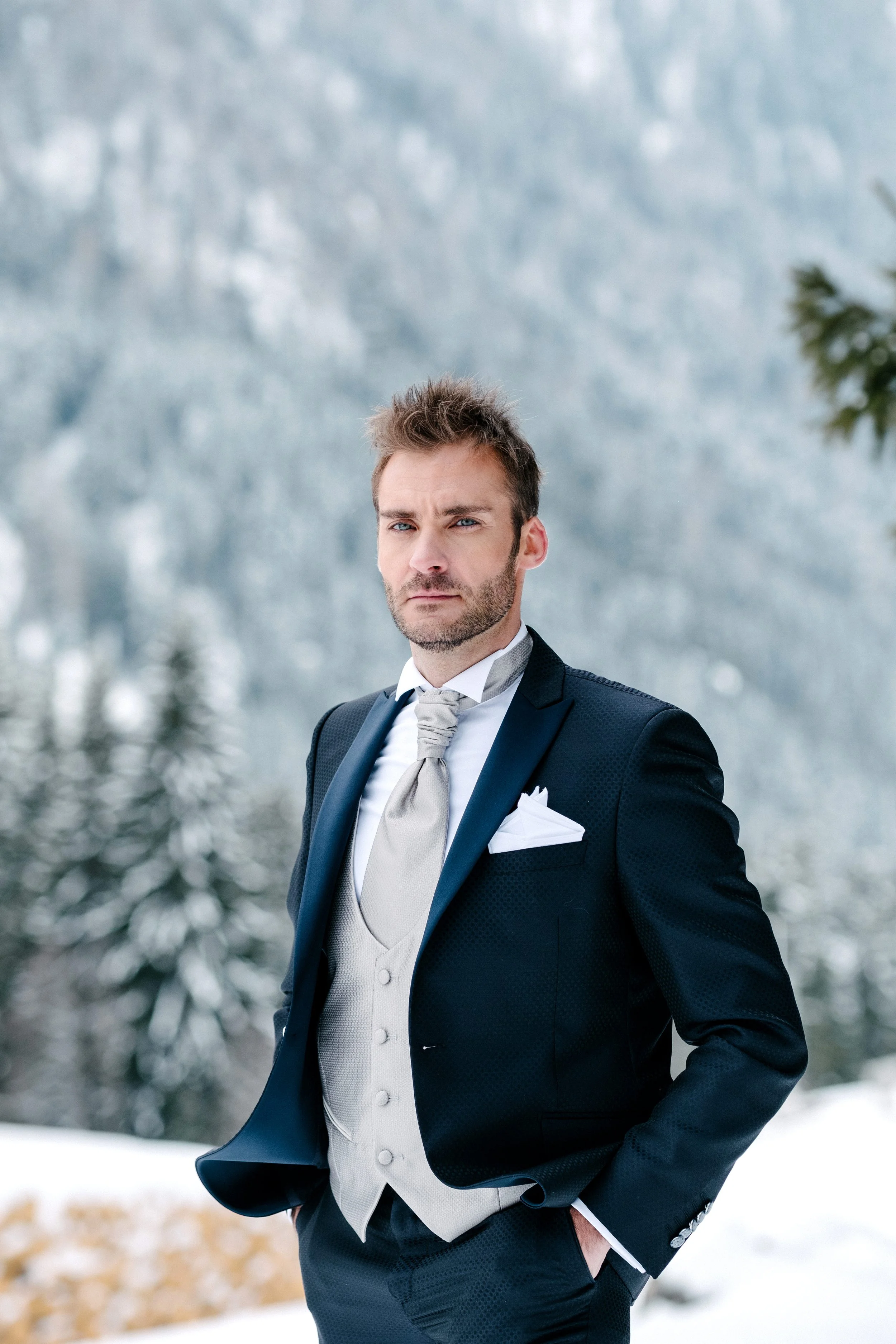A man dressed in a tuxedo standing outdoors in a snowy landscape with trees in the background.