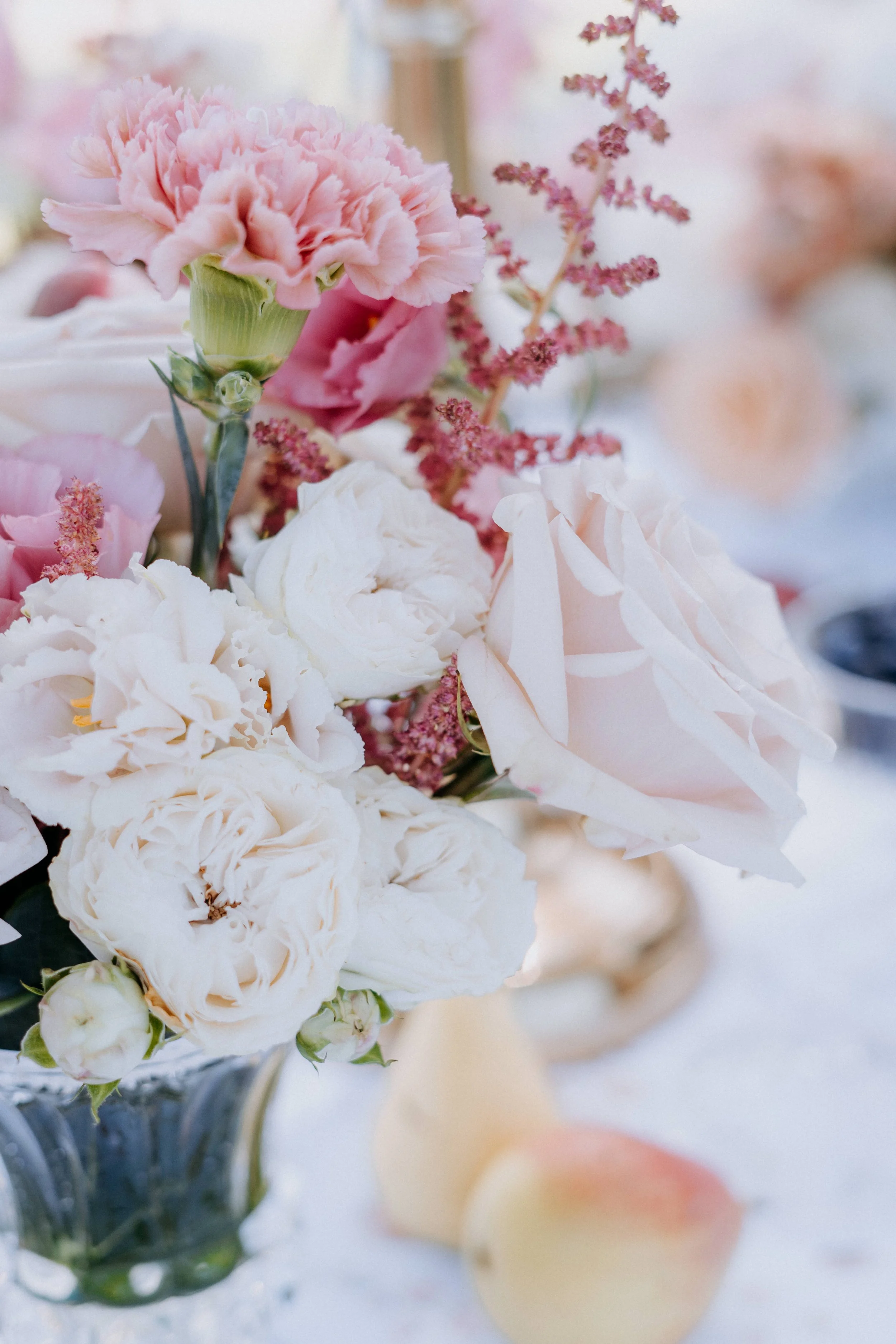 Close-up of a floral arrangement with pink and white carnations, peonies, and roses in a glass vase on a white table.