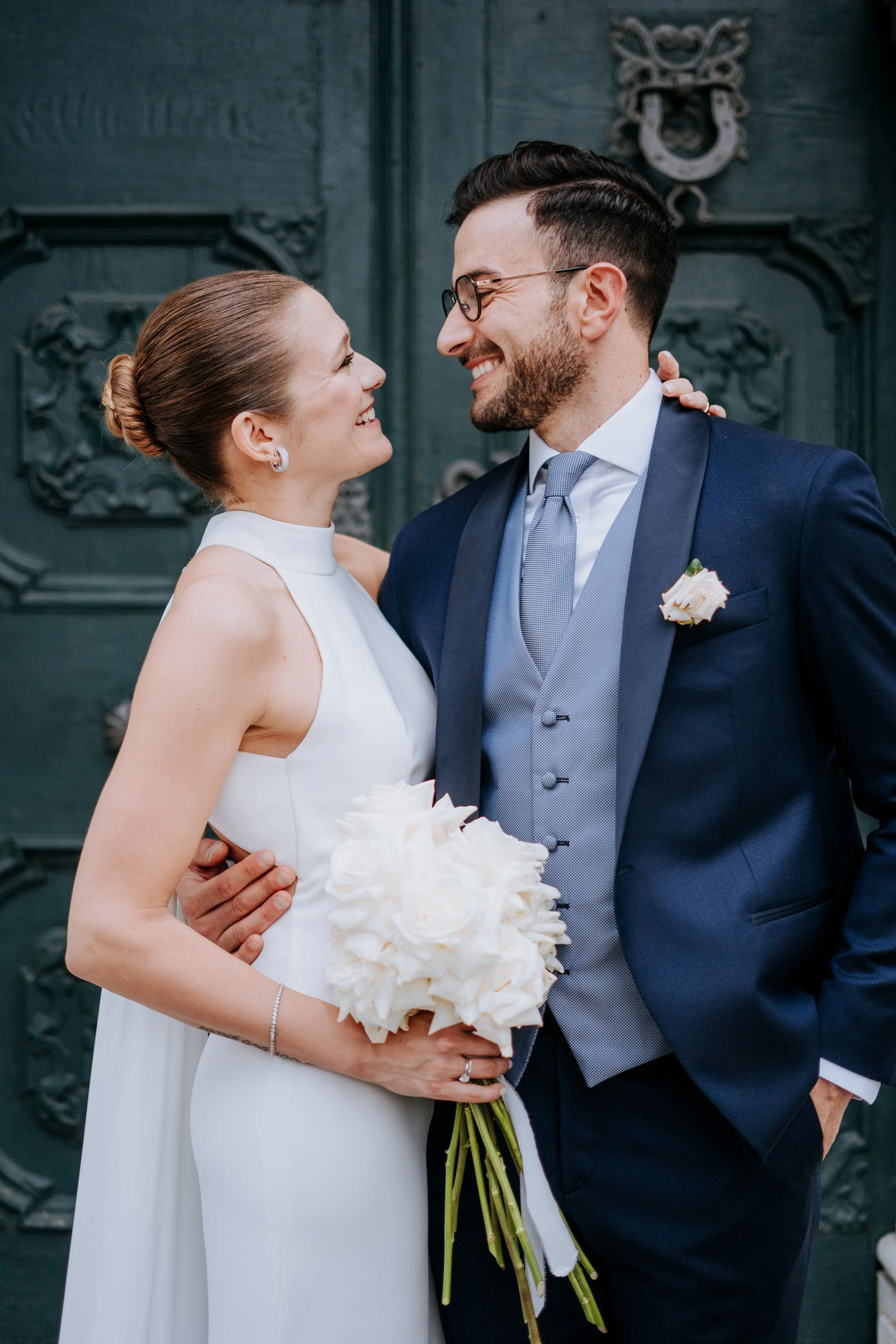 A bride and groom on their wedding day, smiling and looking into each other's eyes, standing in front of an ornate dark door. The bride holds a bouquet of white roses, and the groom wears a navy suit with a gray vest, tie, and a white boutonniere.