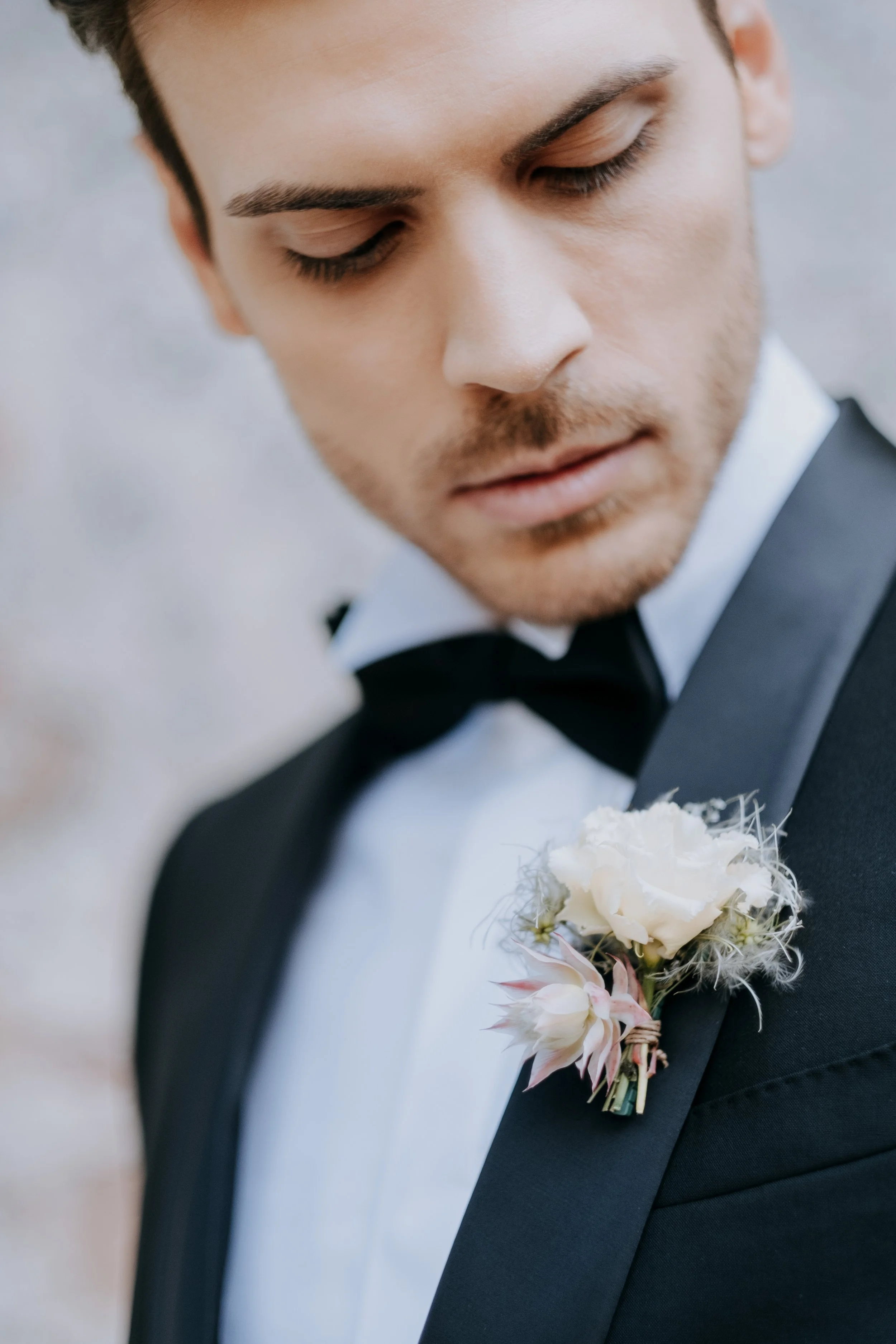 Close-up of a groom in a tuxedo with a black bow tie and white shirt, looking down at a floral boutonniere on his lapel.