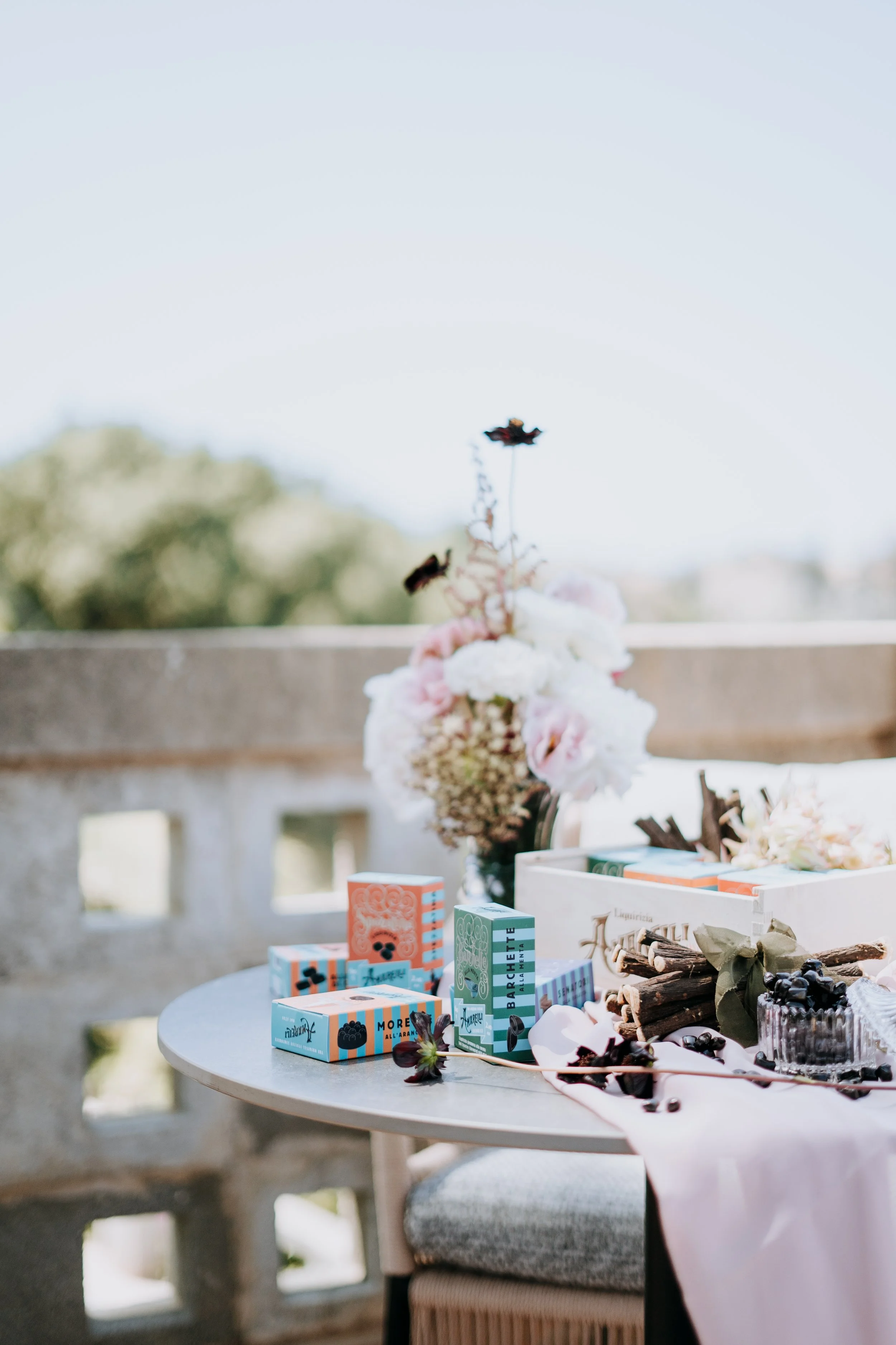 Decorative table with pink roses, dark chocolates, and colorful boxed sparklers, set outdoors near a stone wall.