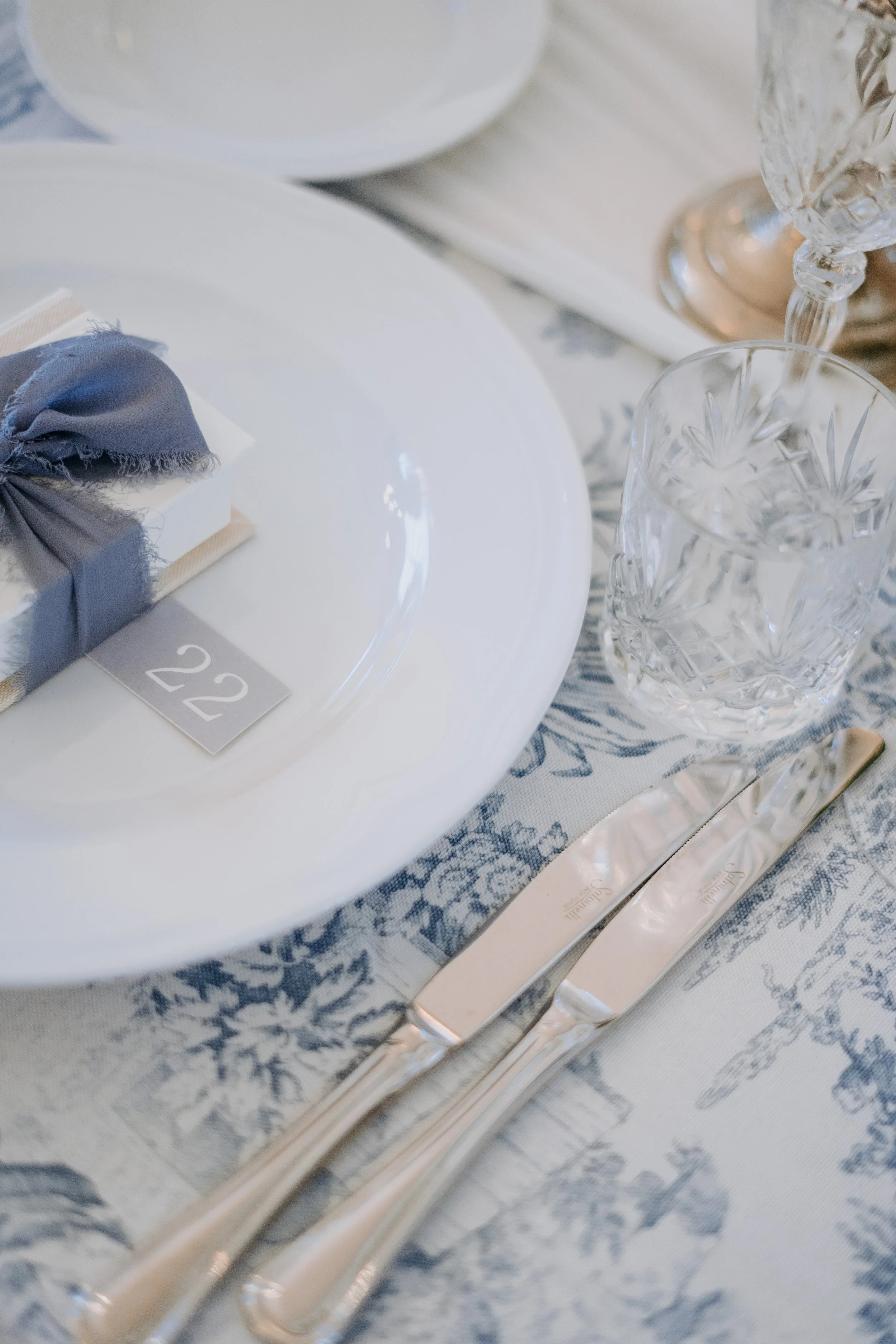 Elegant table setting with a white plate, silverware, crystal glassware, and a blue napkin tied with a gray ribbon, on a patterned tablecloth.
