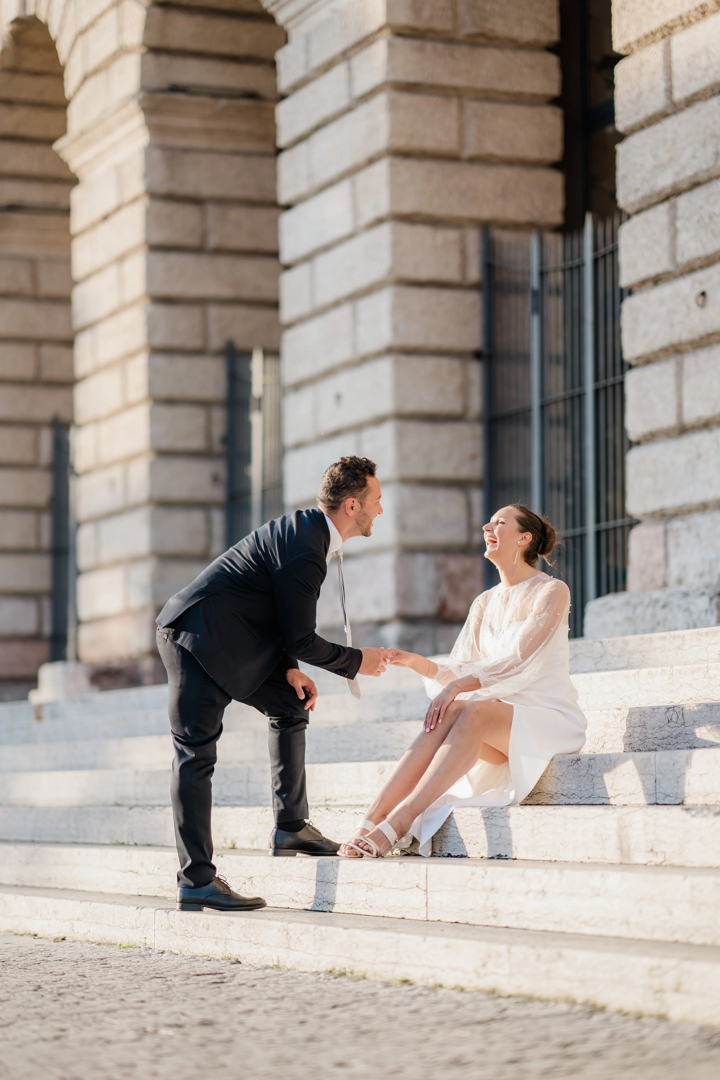 A man in a black suit and woman in white dress sitting on steps outside a building with stone columns, sharing a joyful moment, holding hands and smiling at each other.