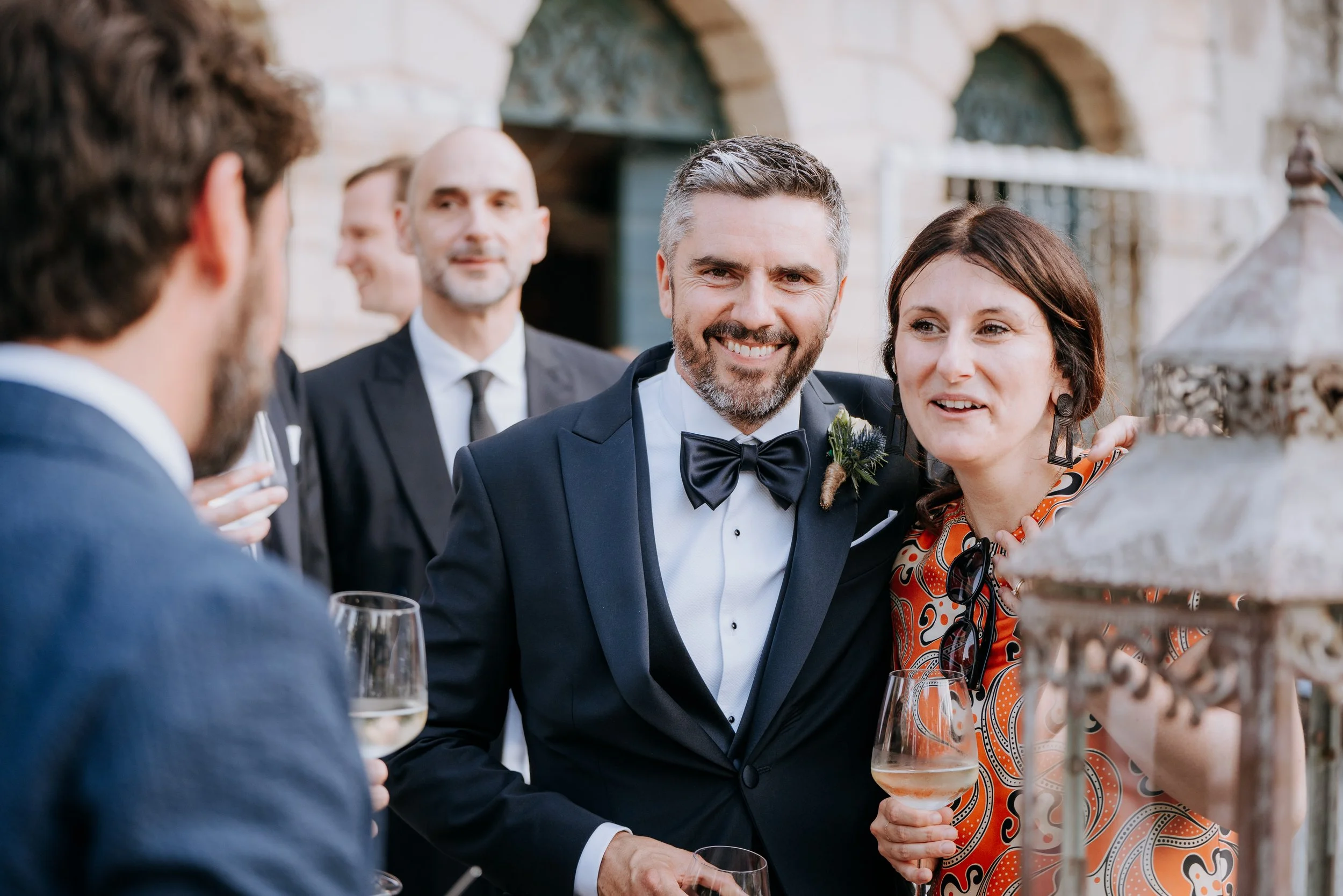 People at a wedding reception, with a man in a tuxedo and a woman with glasses and earrings, talking and holding wine glasses, outside in front of a stone building.