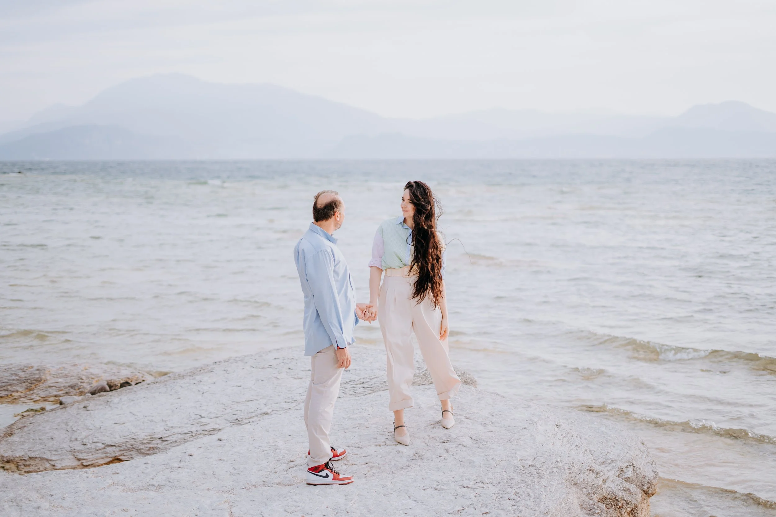 A man and a woman hold hands on a rocky shoreline with the ocean and mountains in the background.