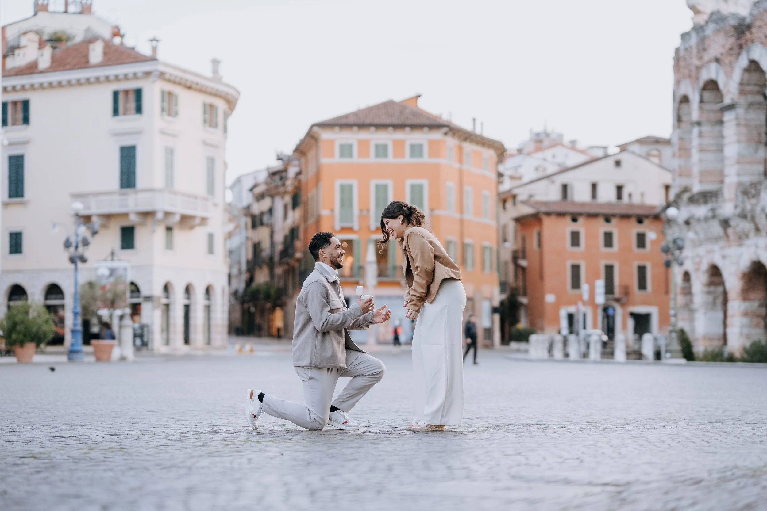 A man proposing marriage to a woman in a European city square with colorful old buildings in the background.