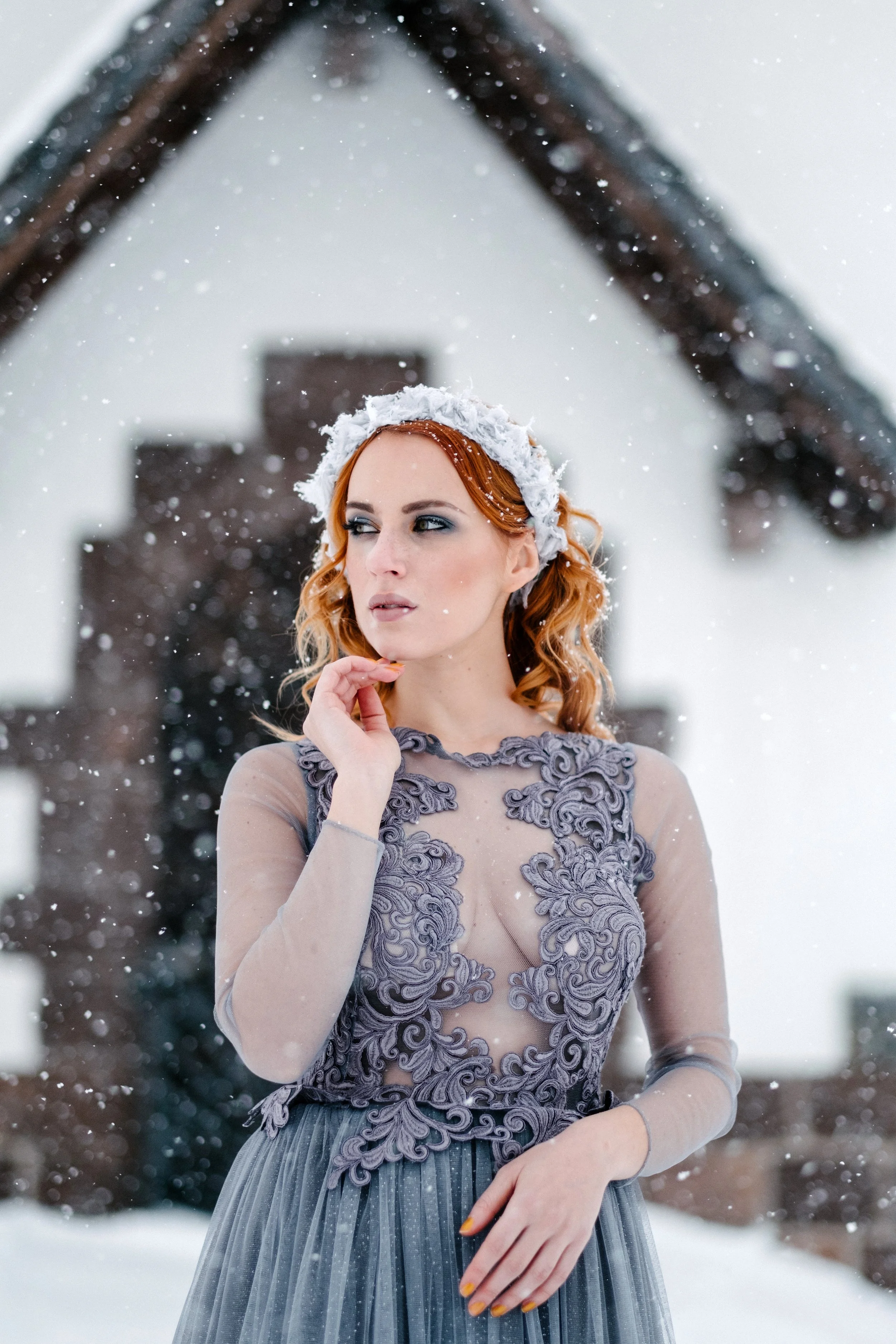A woman with red hair wearing a sheer, embroidered dress and a white, fluffy headpiece stands in the snow with a blurred house in the background.