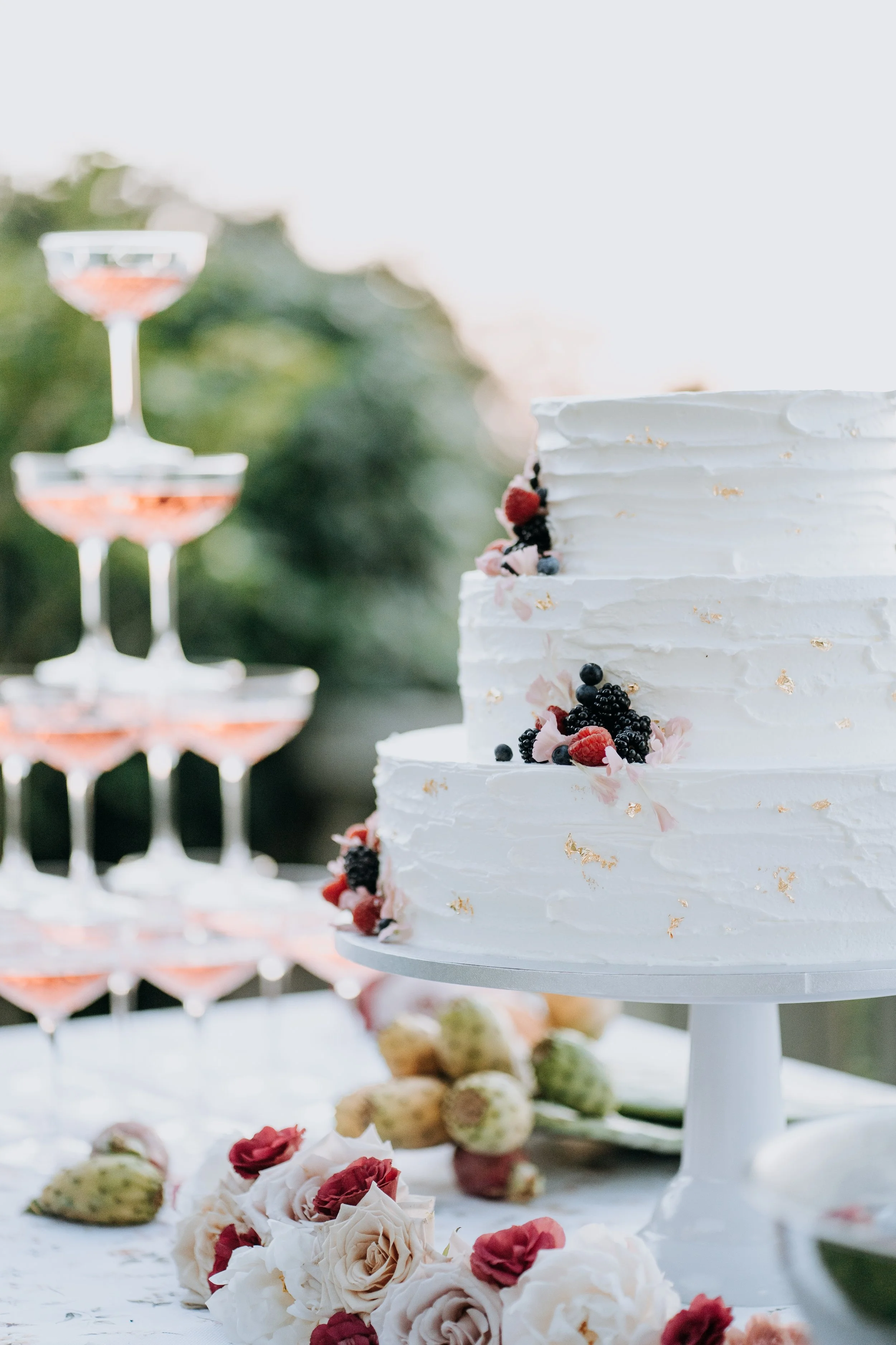 A white two-tier wedding cake decorated with berries and pink flowers, displayed on a cake stand, with a dessert tower of wine glasses filled with a pink drink in the background, outdoors with greenery.
