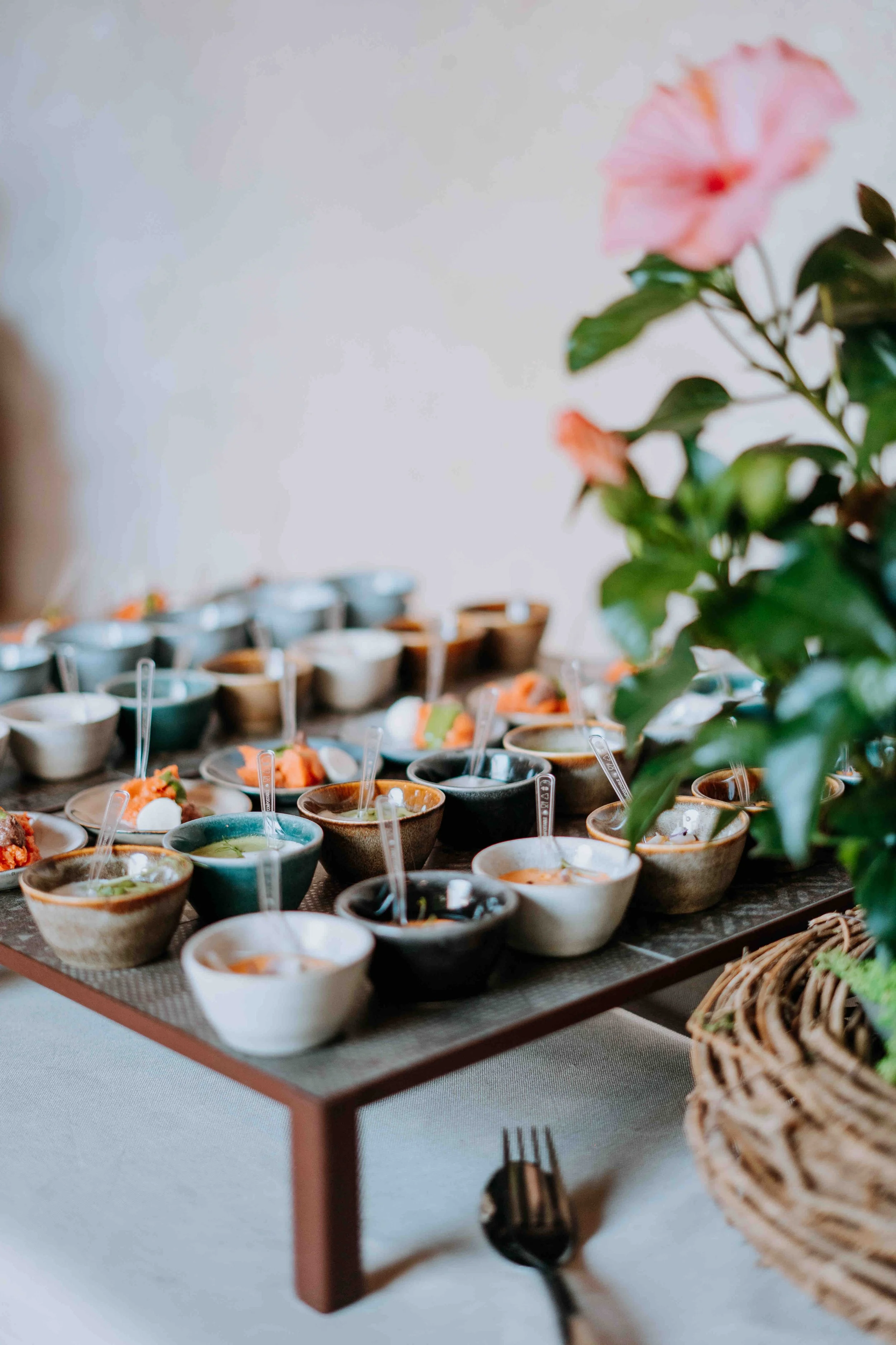 A wooden tray with numerous small bowls filled with various sauces and condiments, some with small tasting spoons, on a table near a basket and a plant with pink flowers.