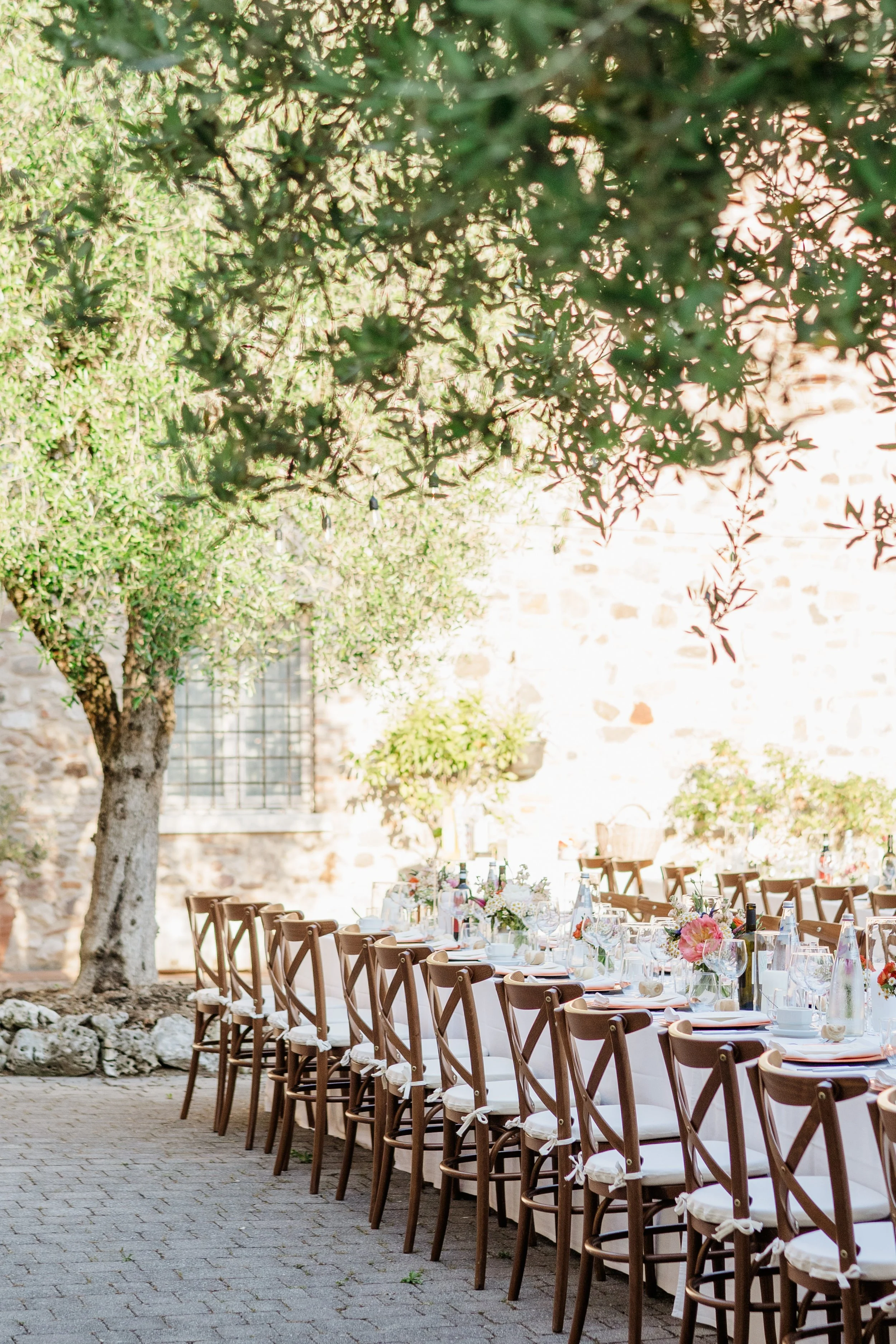 A long outdoor dining table set with white tablecloths, glasses, bottles, and floral centerpieces, situated in a garden area with a large tree and a stone building in the background.