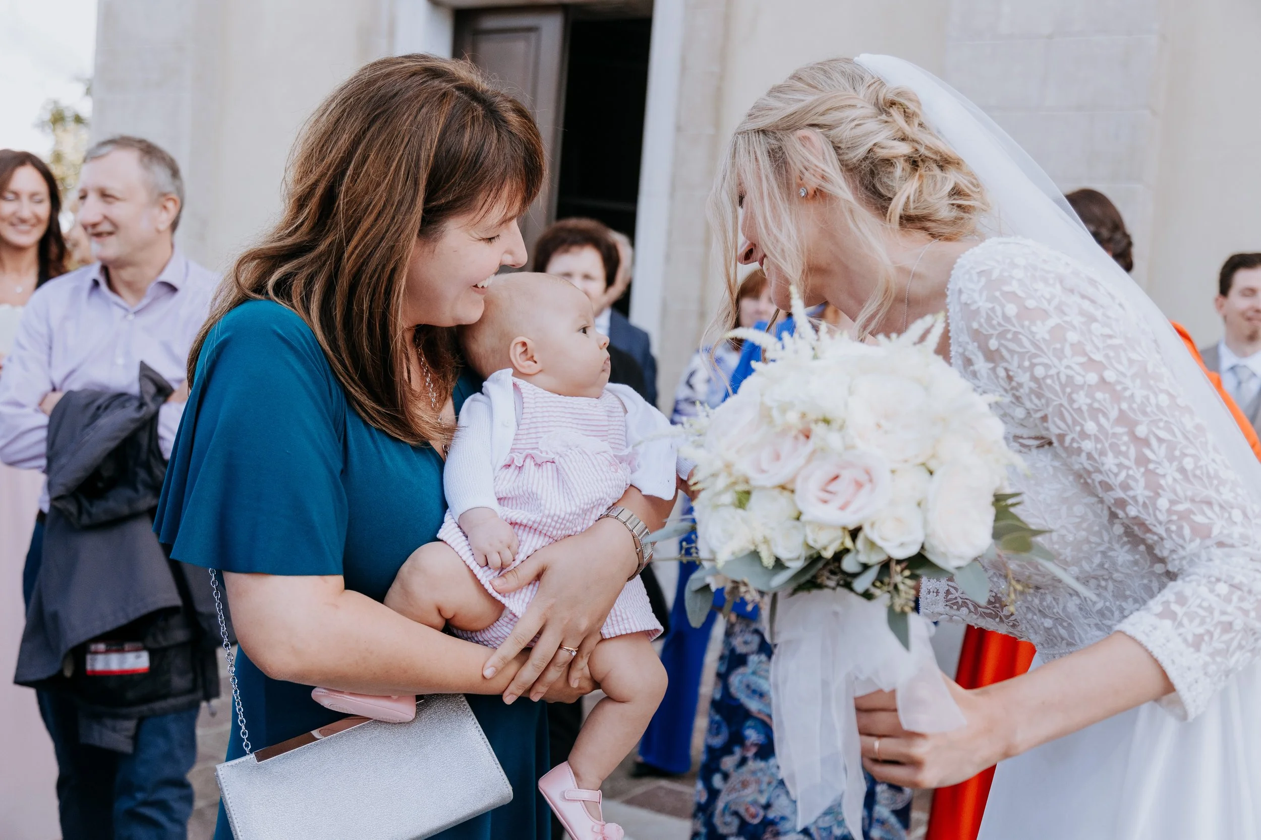 Bride in white lace dress leaning down to look at a baby being held by a woman in blue at a wedding, holding a bouquet of white and pink roses while other guests smile in the background.