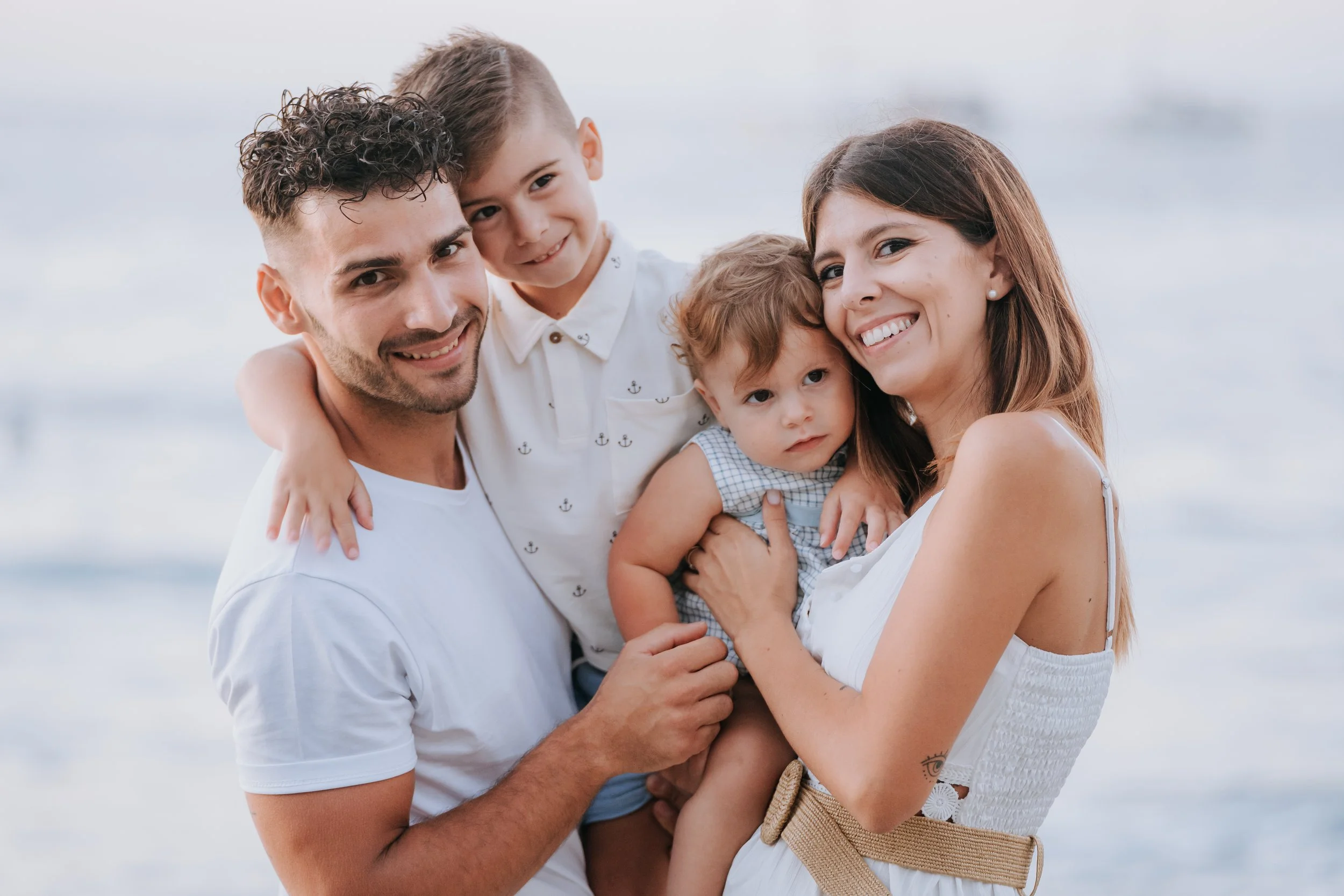 A happy family of four smiling at the camera at the beach with the ocean in the background. The father is holding a young boy, the mother is holding a younger girl, and they are all dressed in casual summer clothes.