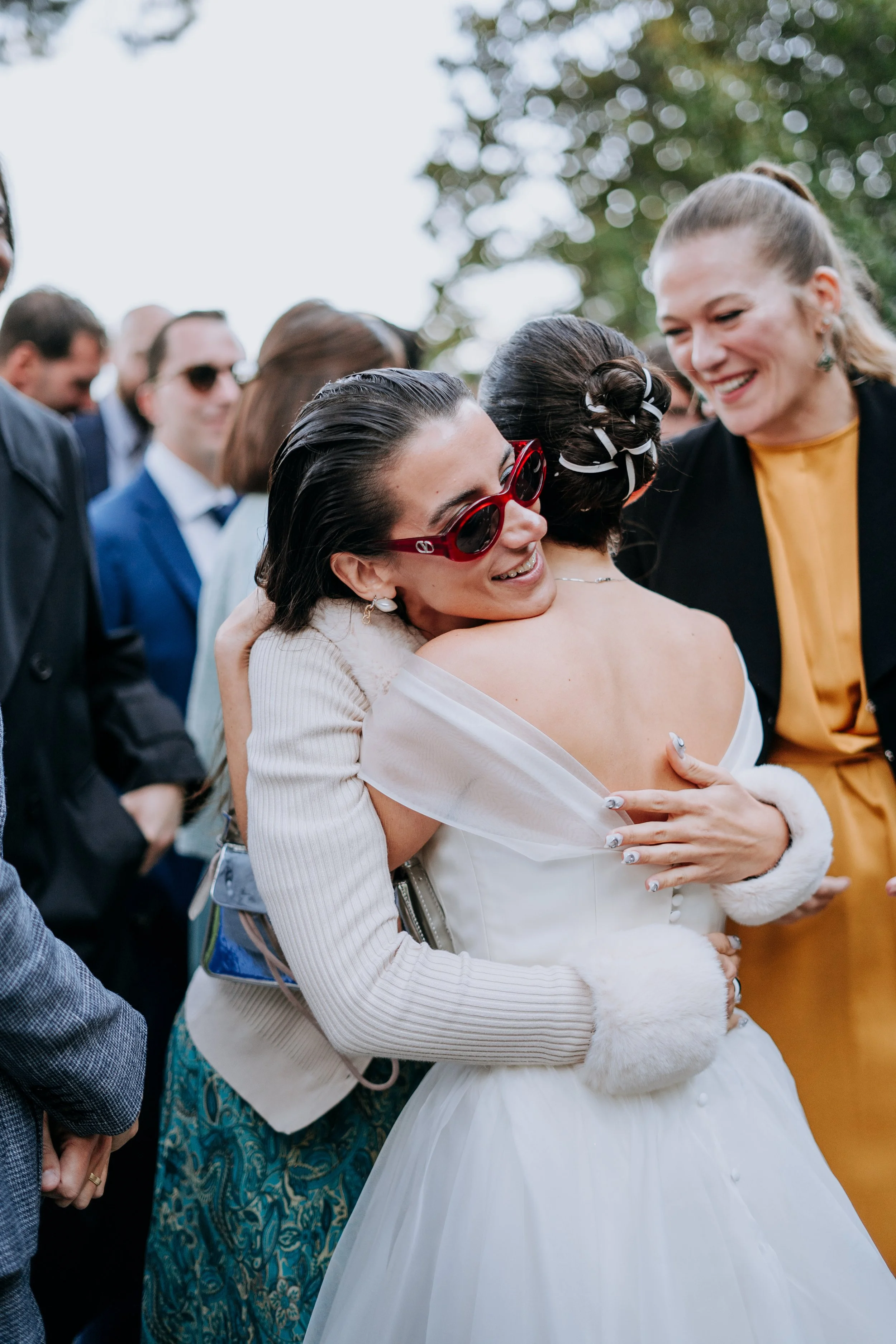 Two women hugging at a wedding, one in a white dress and the other wearing red sunglasses and a cream-colored jacket, while other guests are smiling and talking in the background.