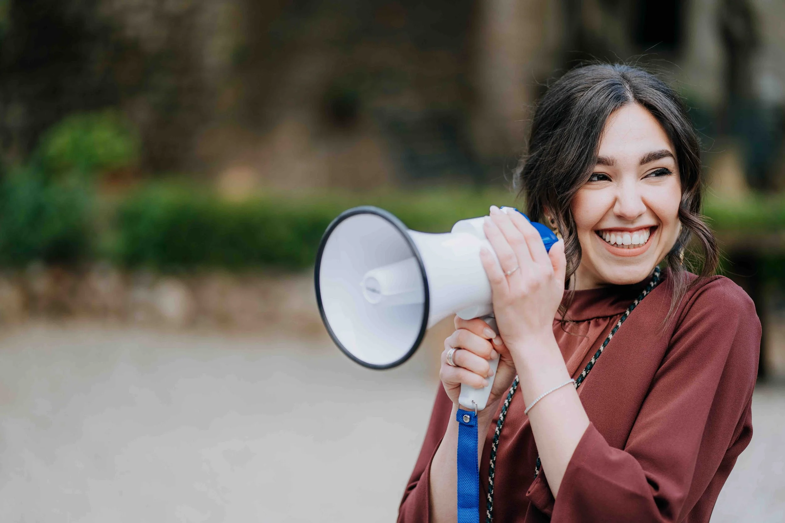 A young woman with brown hair smiling while holding a white megaphone outdoors.