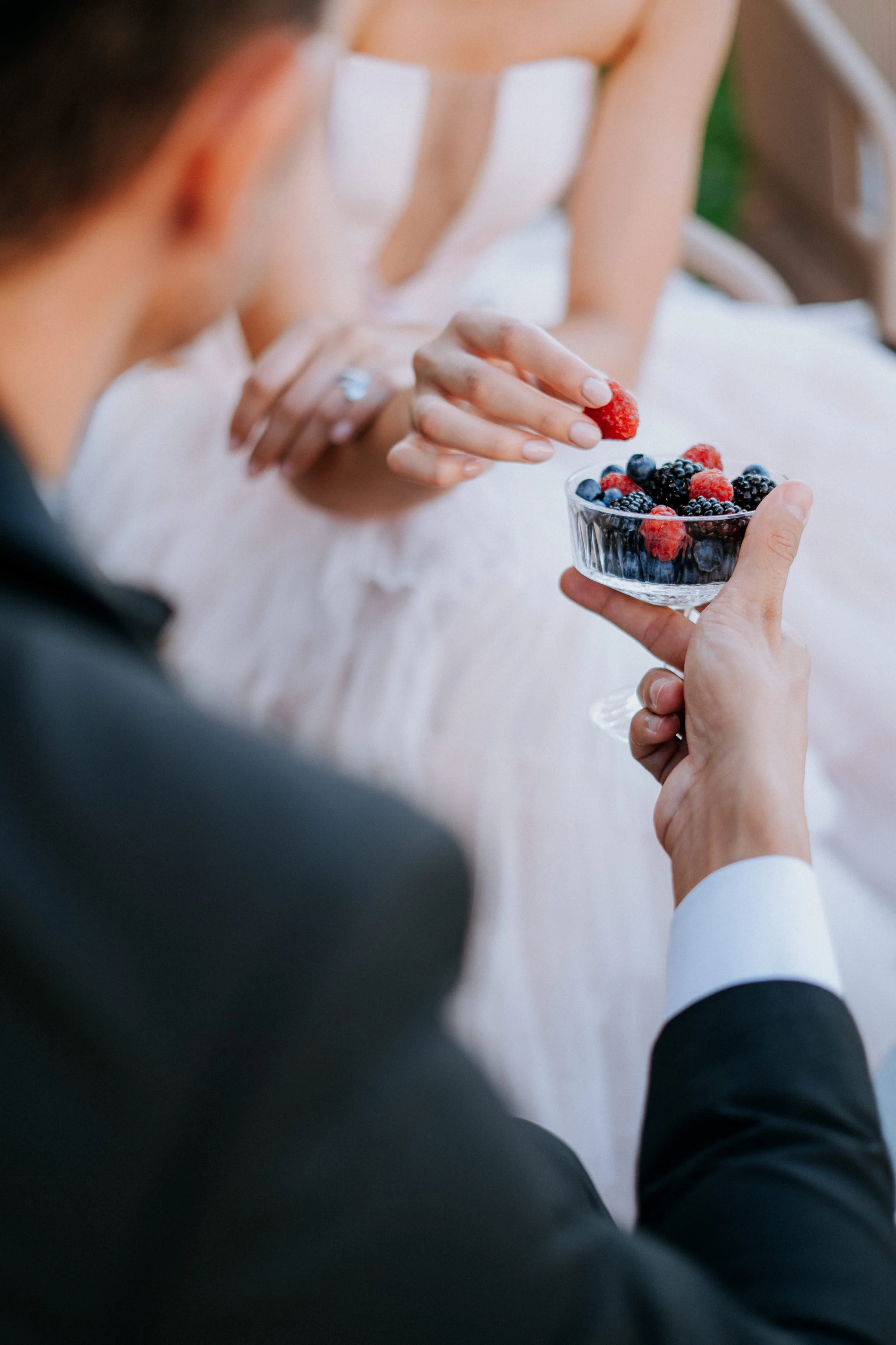 A man in a tuxedo is holding a glass bowl of mixed berries, including raspberries, blackberries, and blueberries, while a woman in a wedding dress reaches to pick a berry from the bowl.