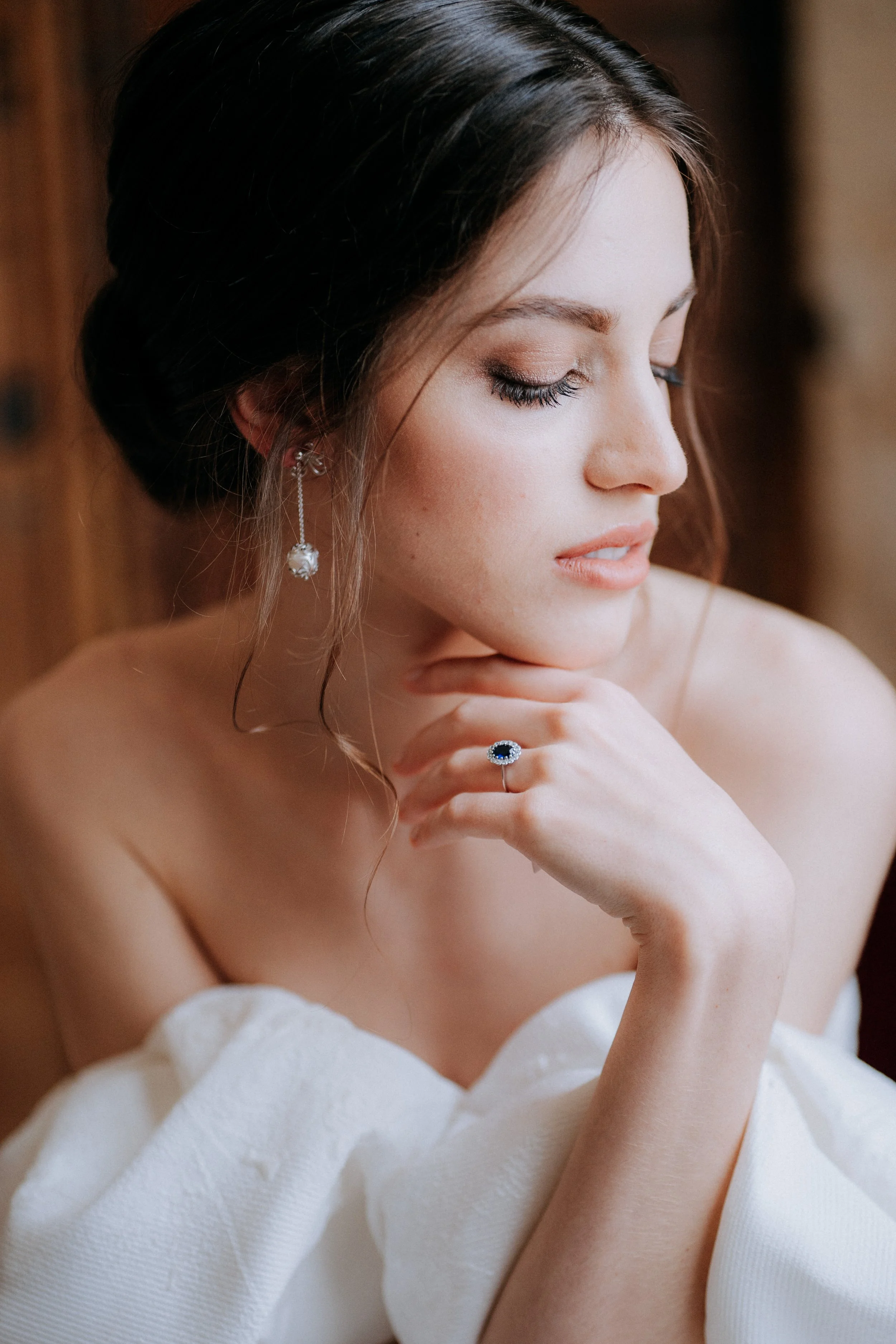 A woman with dark hair styled in an elegant updo, wearing a white off-the-shoulder garment, appears to be in a contemplative pose with her eyes closed and her hand resting on her chin. She is adorned with a pair of dangling earrings and a large ring