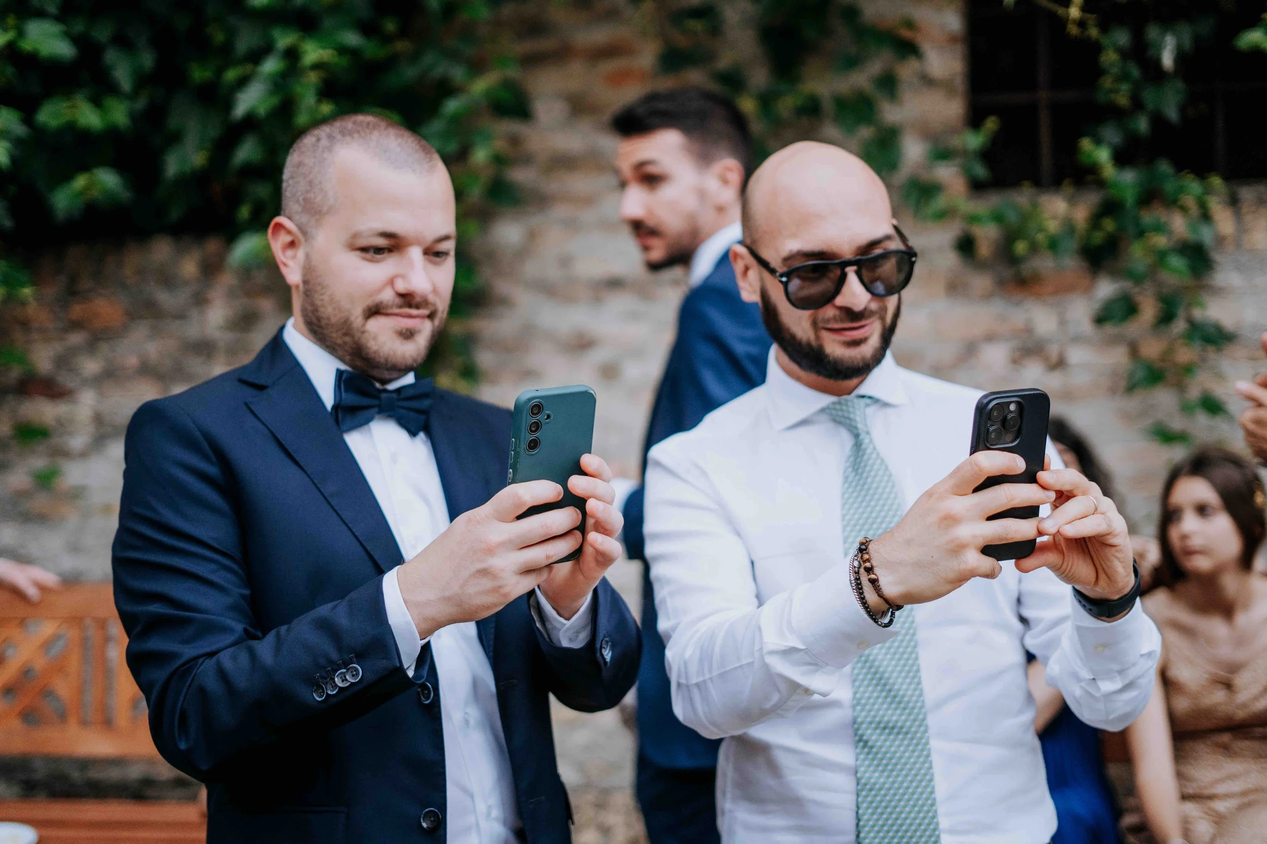 Two men dressed in formal suits and ties are taking photos or videos with their smartphones at an outdoor event. Other people are visible in the background, and a brick wall with greenery is behind them.