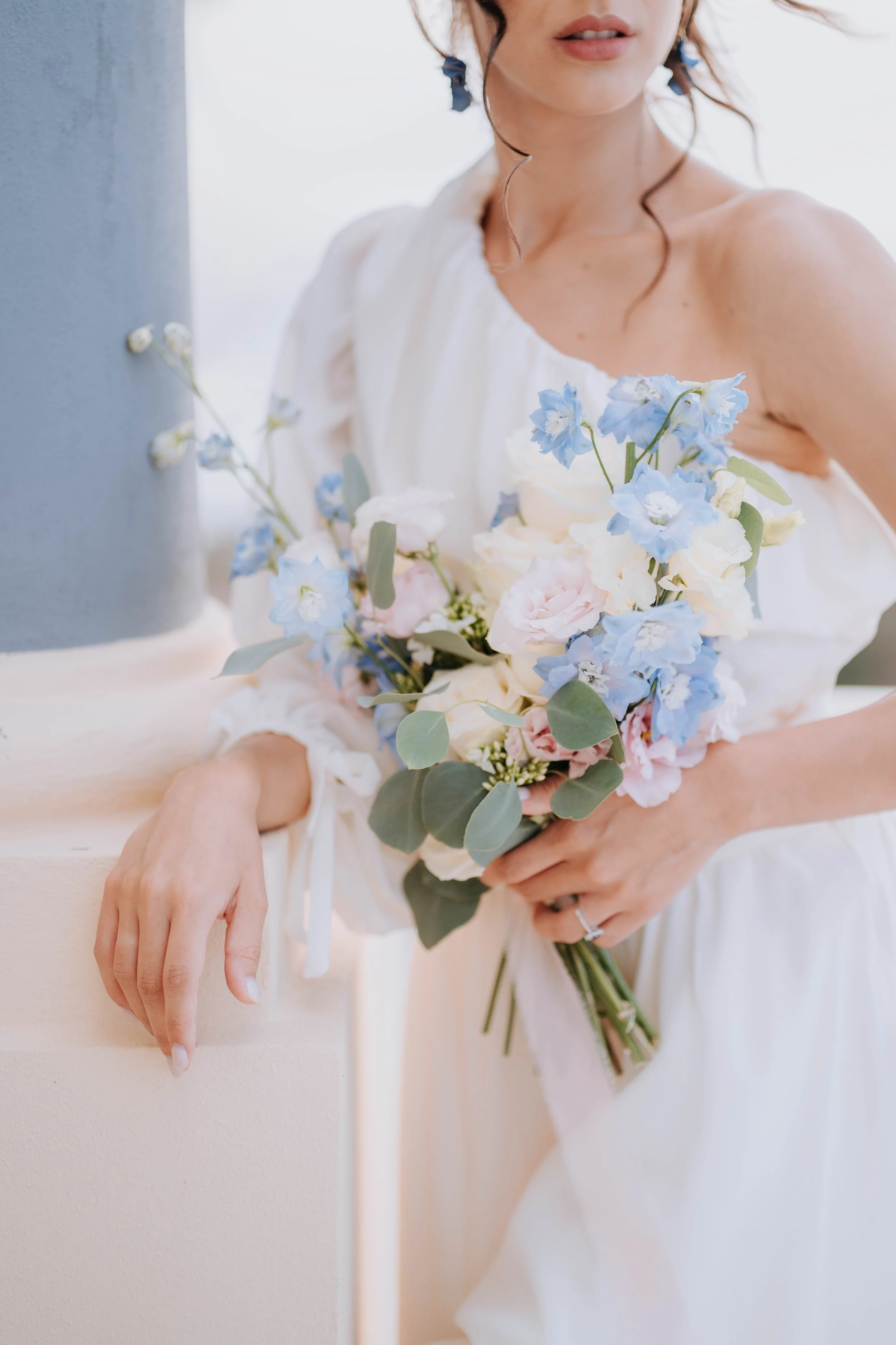 A woman in a white dress holding a pastel-colored bouquet of flowers, with part of her face and shoulder visible, against a light background.