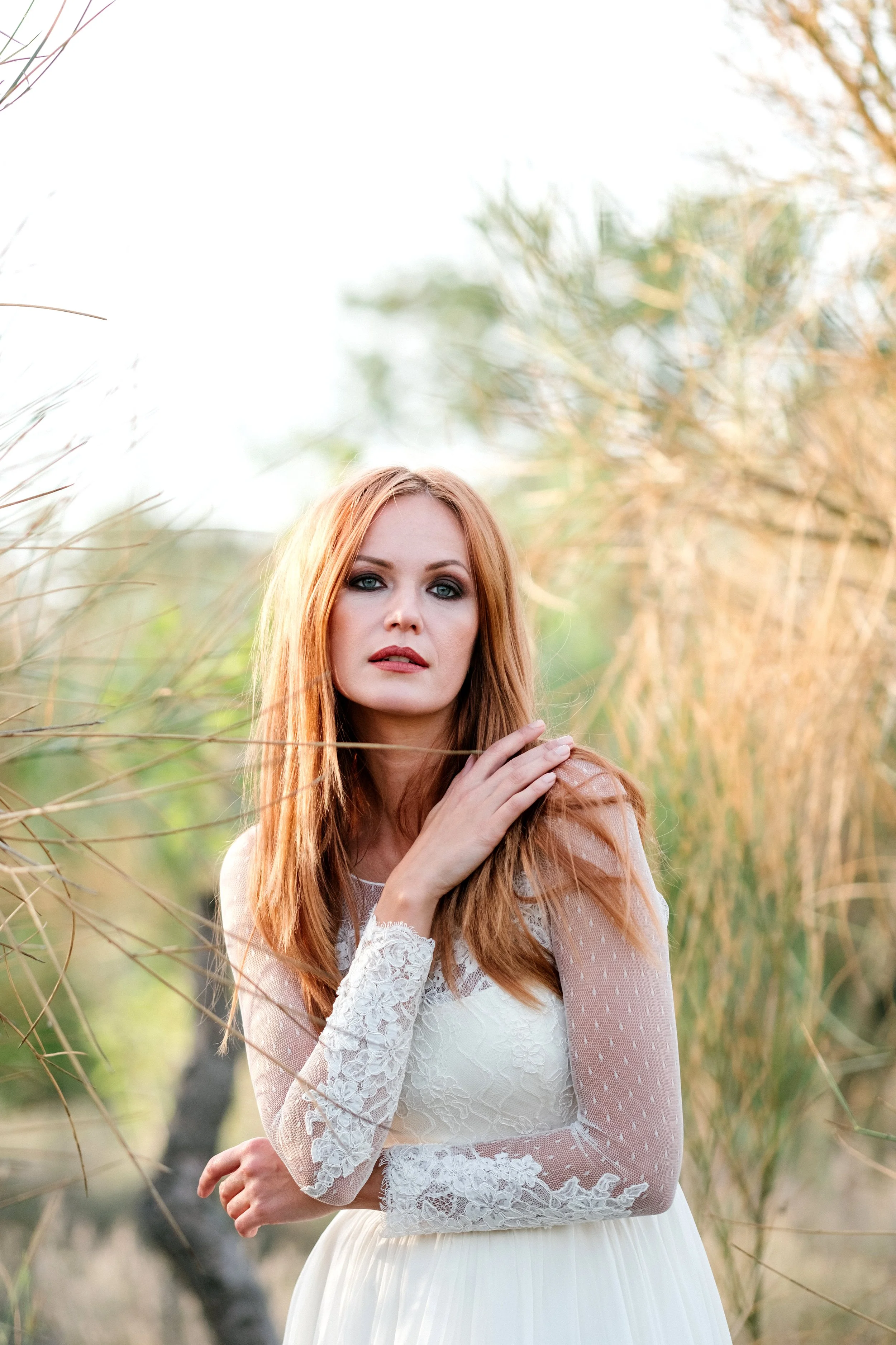 A woman with red hair in a white lace dress stands outdoors among dry grass and trees, looking at the camera.