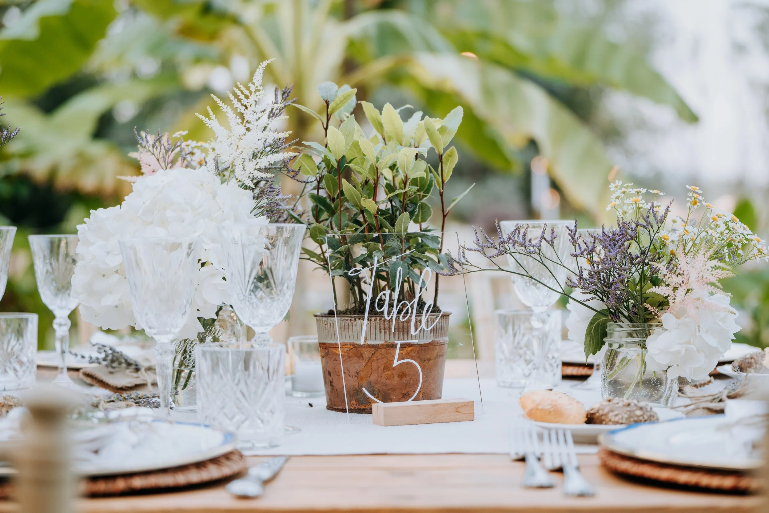 Table decorated with white floral arrangements, green foliage, and glassware, with a centerpiece sign that says 'Table 5' at an outdoor event.