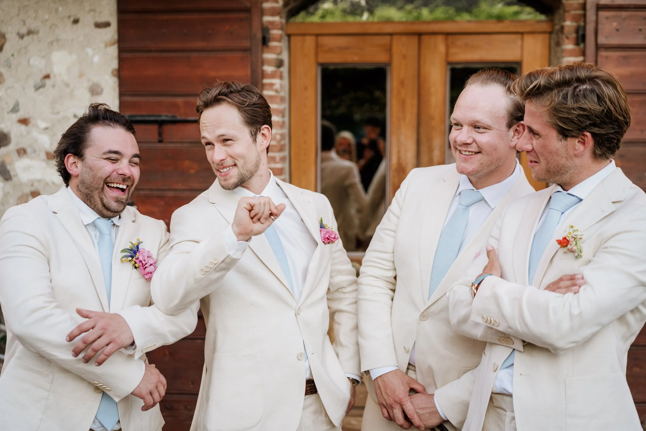 Four men dressed in white suits and light blue ties, standing together outdoors in front of a wooden building, sharing a laugh and conversation. One man is gesturing with a fist bump.