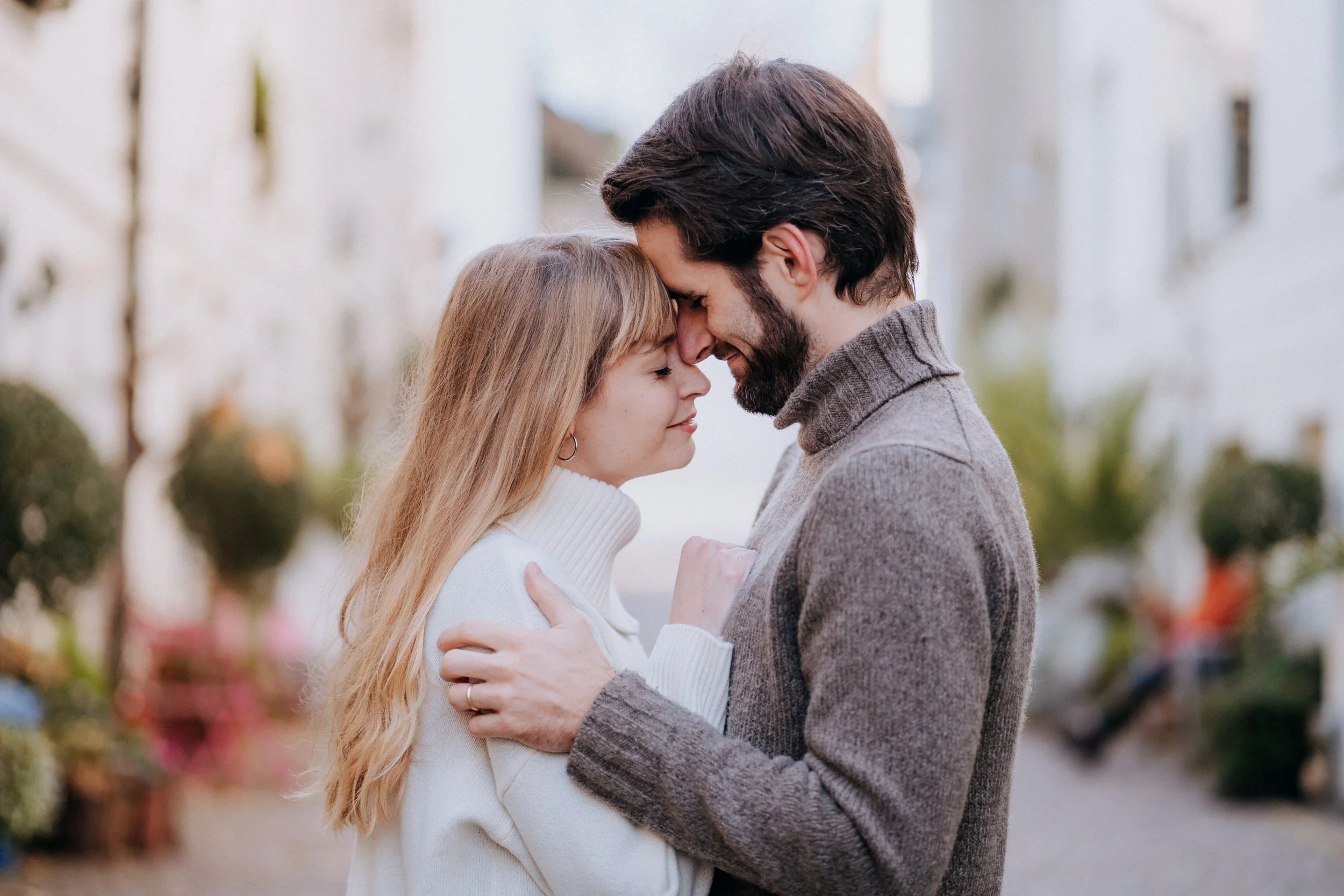 A couple embracing outdoors with their foreheads and noses touching, smiling with their eyes closed.