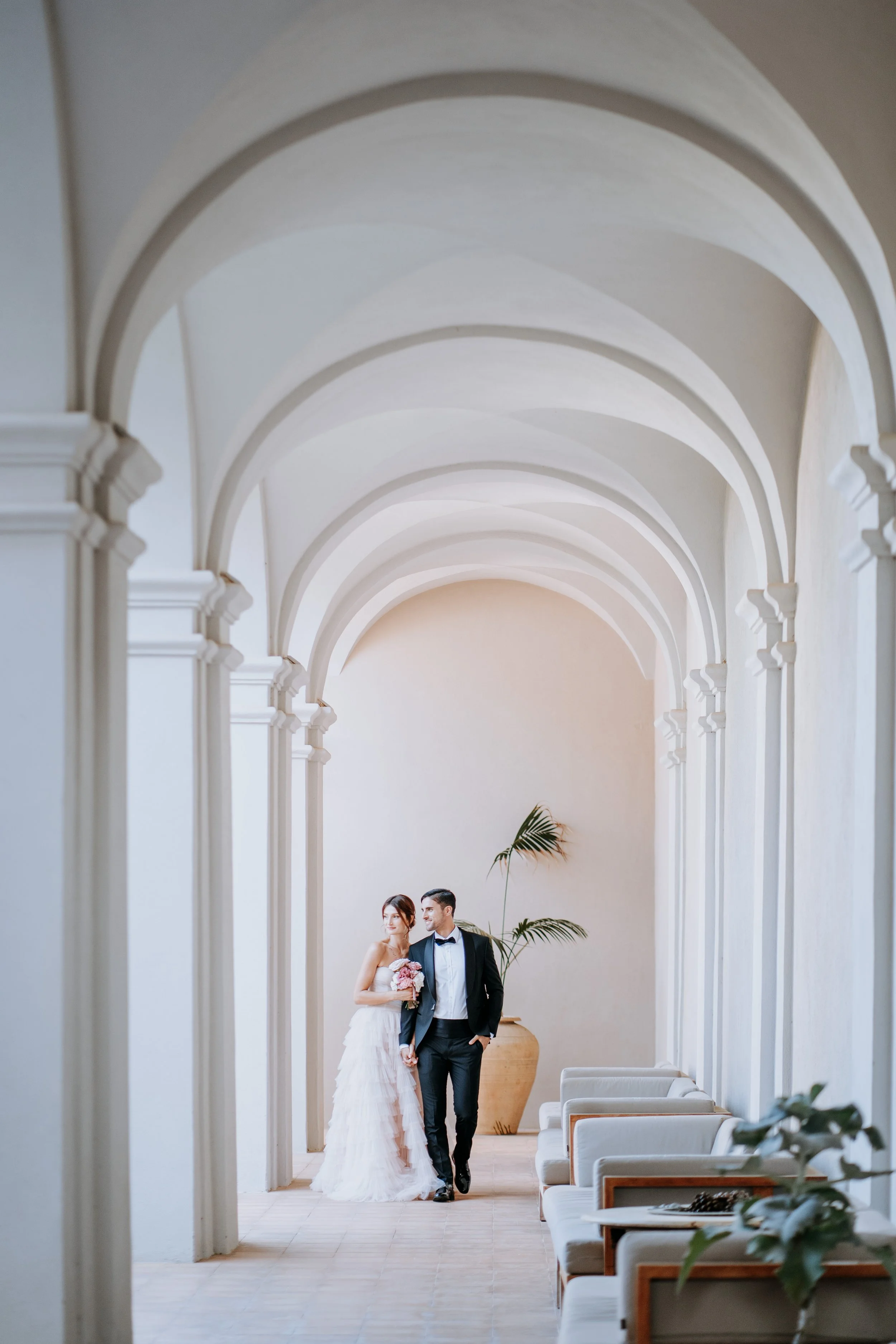 Bride and groom walking in a bright, arched corridor with white walls and chairs on the side, holding hands and looking at each other, with a potted plant in the background.