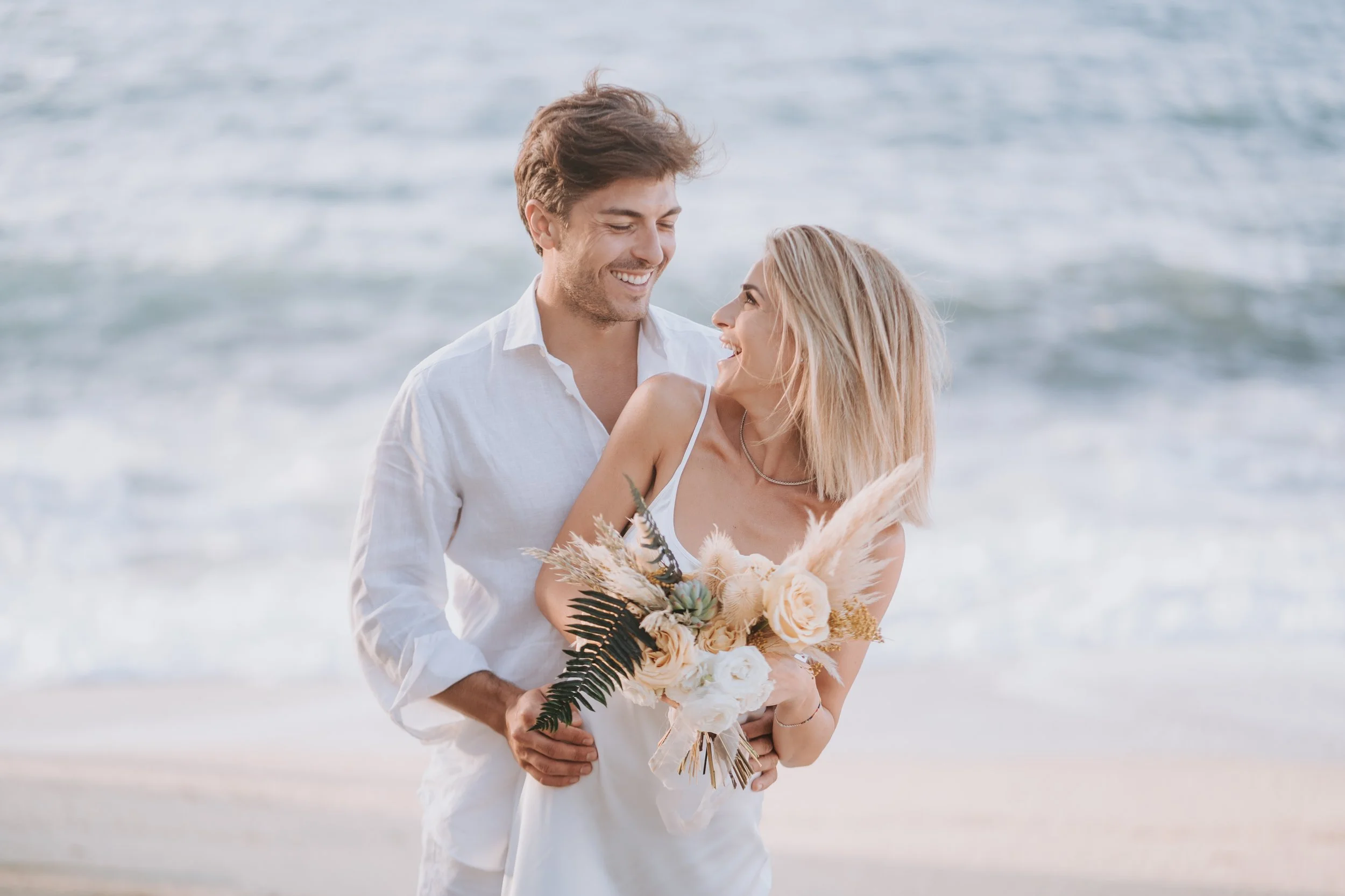A couple dressed in white, smiling and embracing on a beach, with the woman holding a bouquet of flowers.