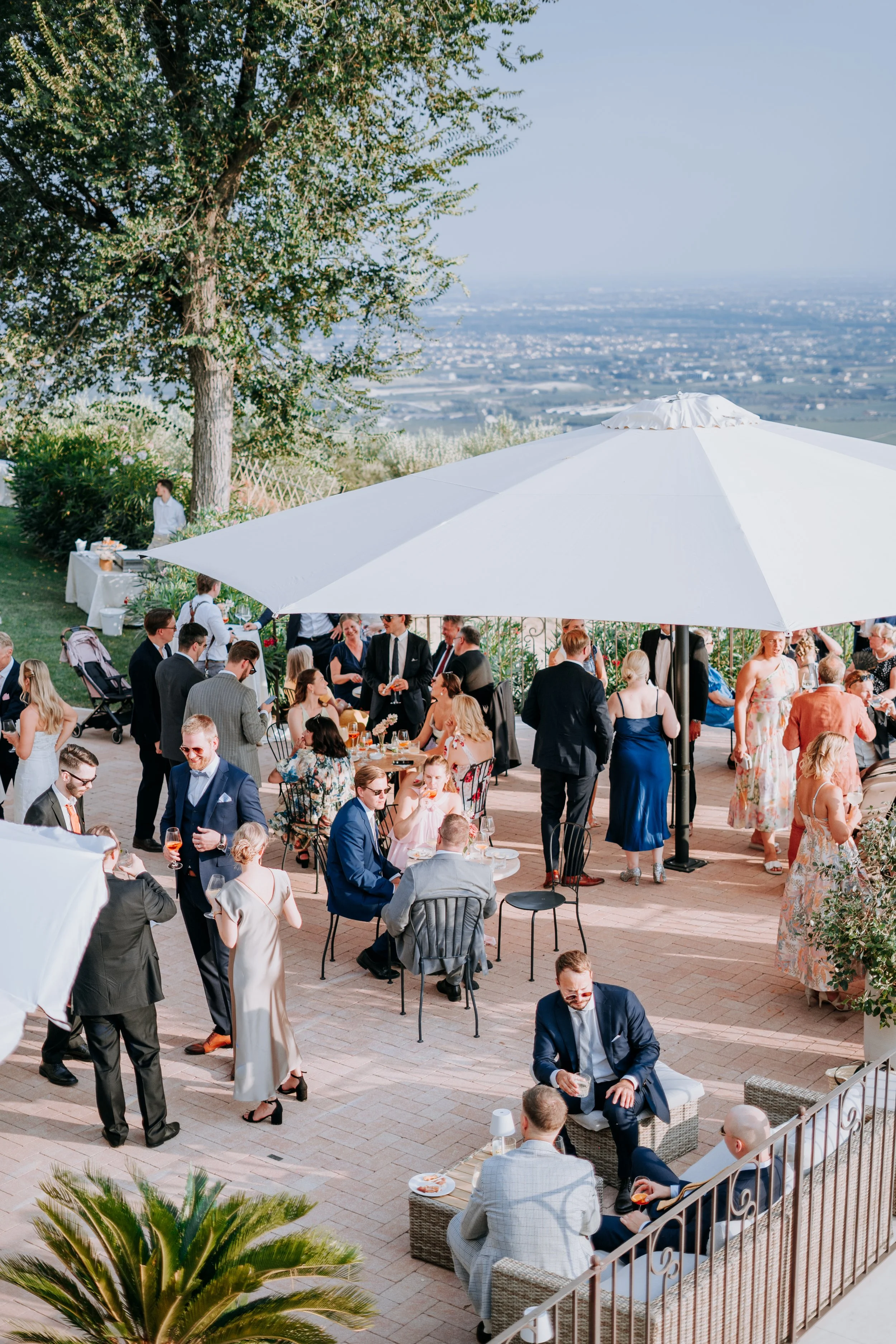 Guests at an outdoor wedding reception under a large white umbrella, with some seated at tables and others mingling, overlooking a scenic view of a valley.