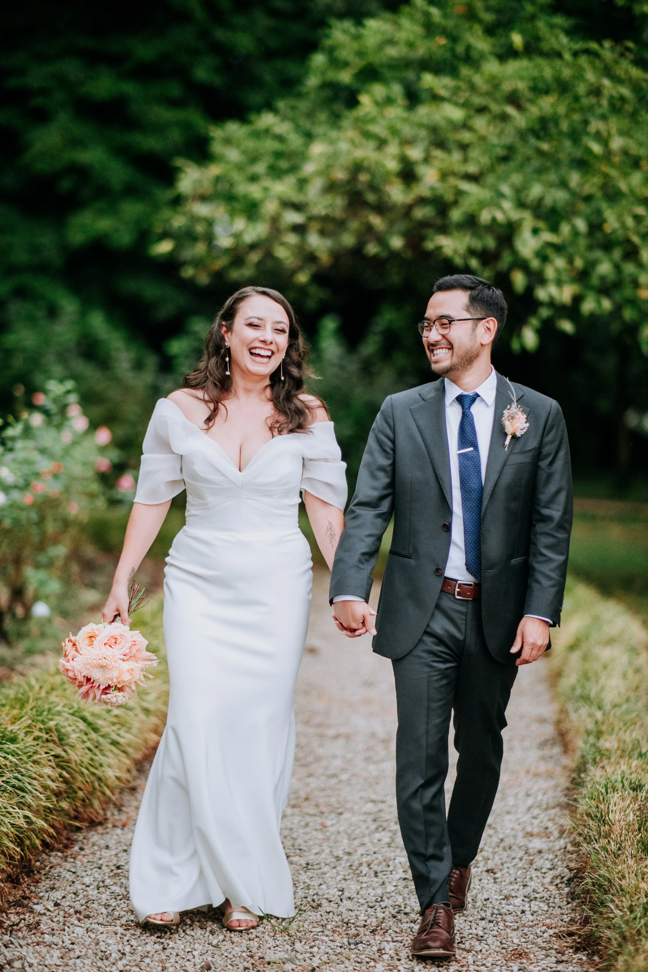 A smiling bride and groom walking hand in hand outdoors, surrounded by greenery, during a wedding celebration.