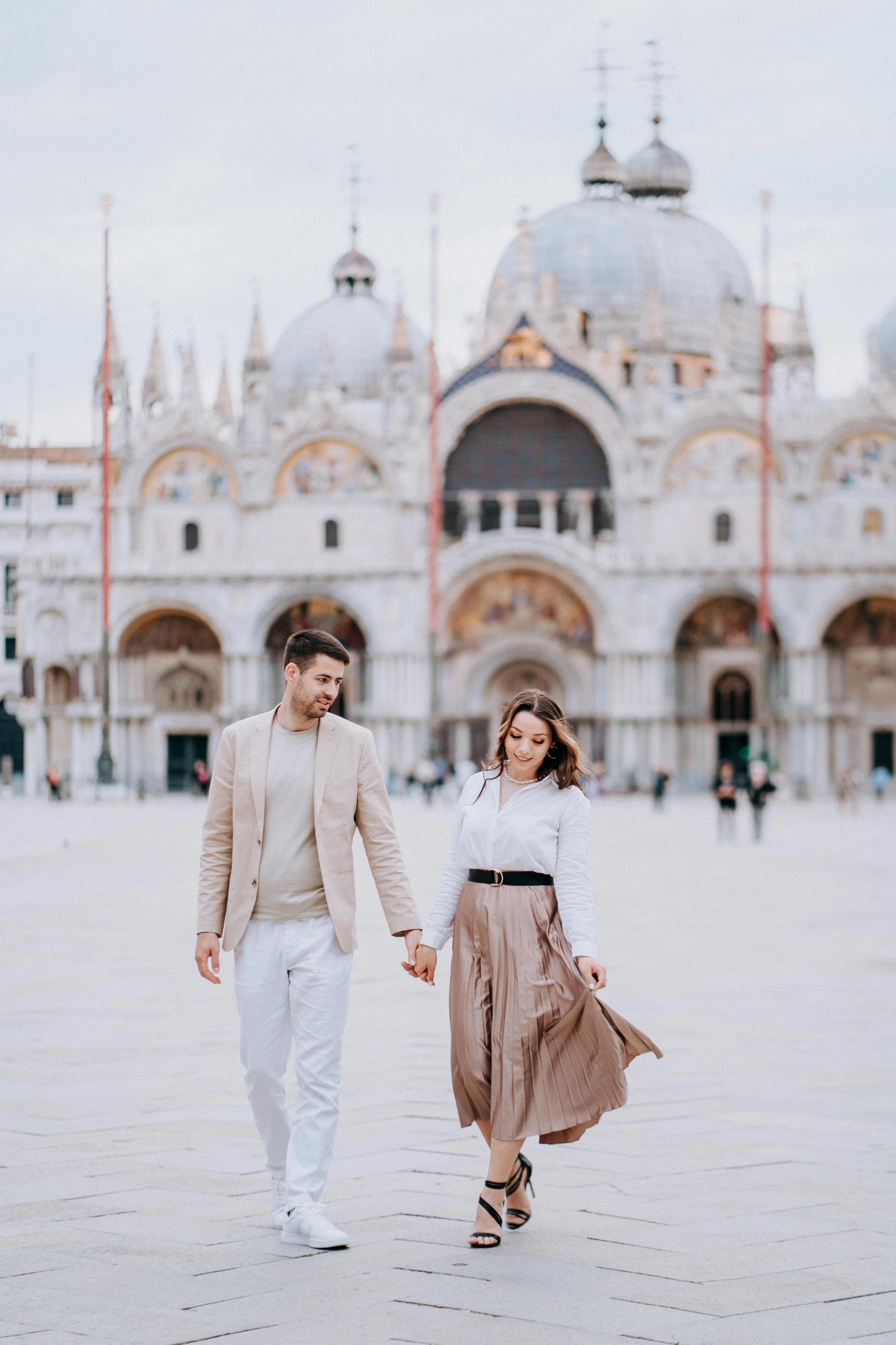 A couple holding hands and walking in front of a historic building with domed towers, possibly St. Mark's Basilica in Venice, Italy.