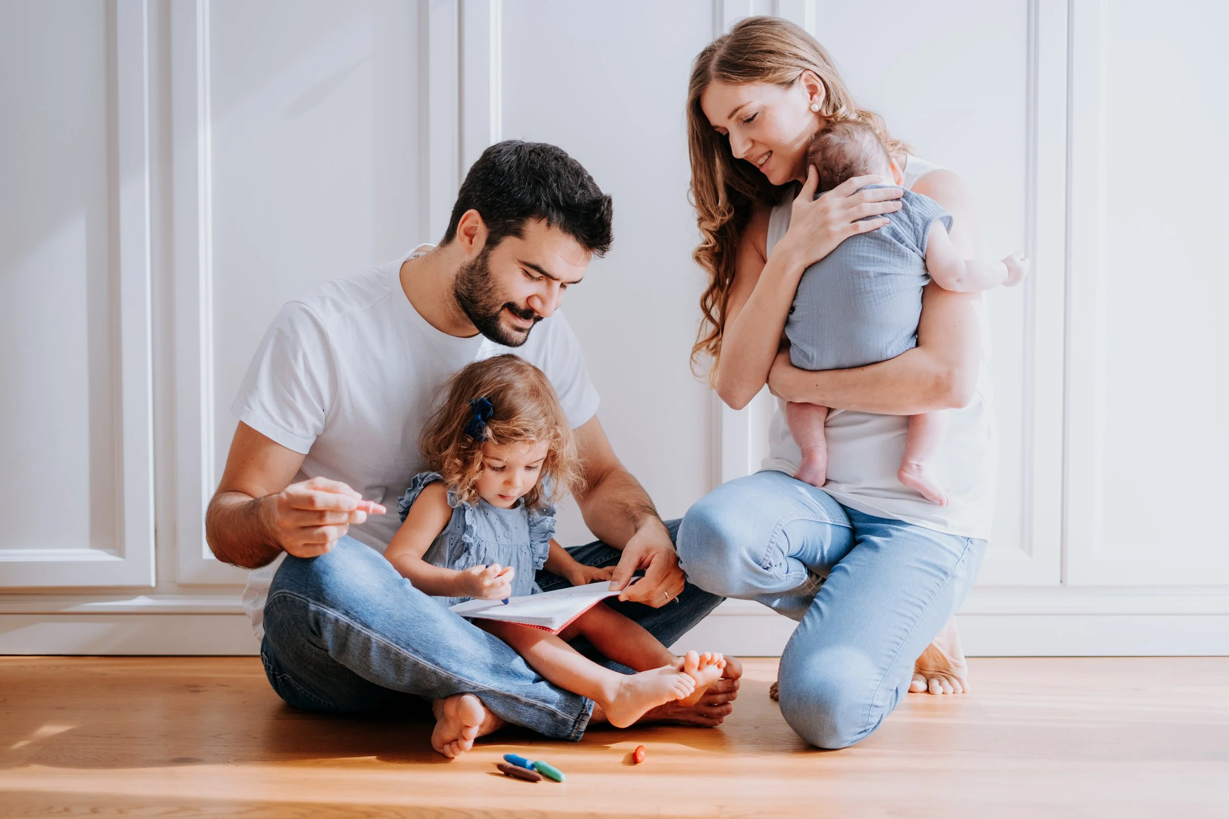 Family of four sitting on hardwood floor, with mother holding a baby, father and daughter looking at a book. Crayons scattered nearby, bright natural light.