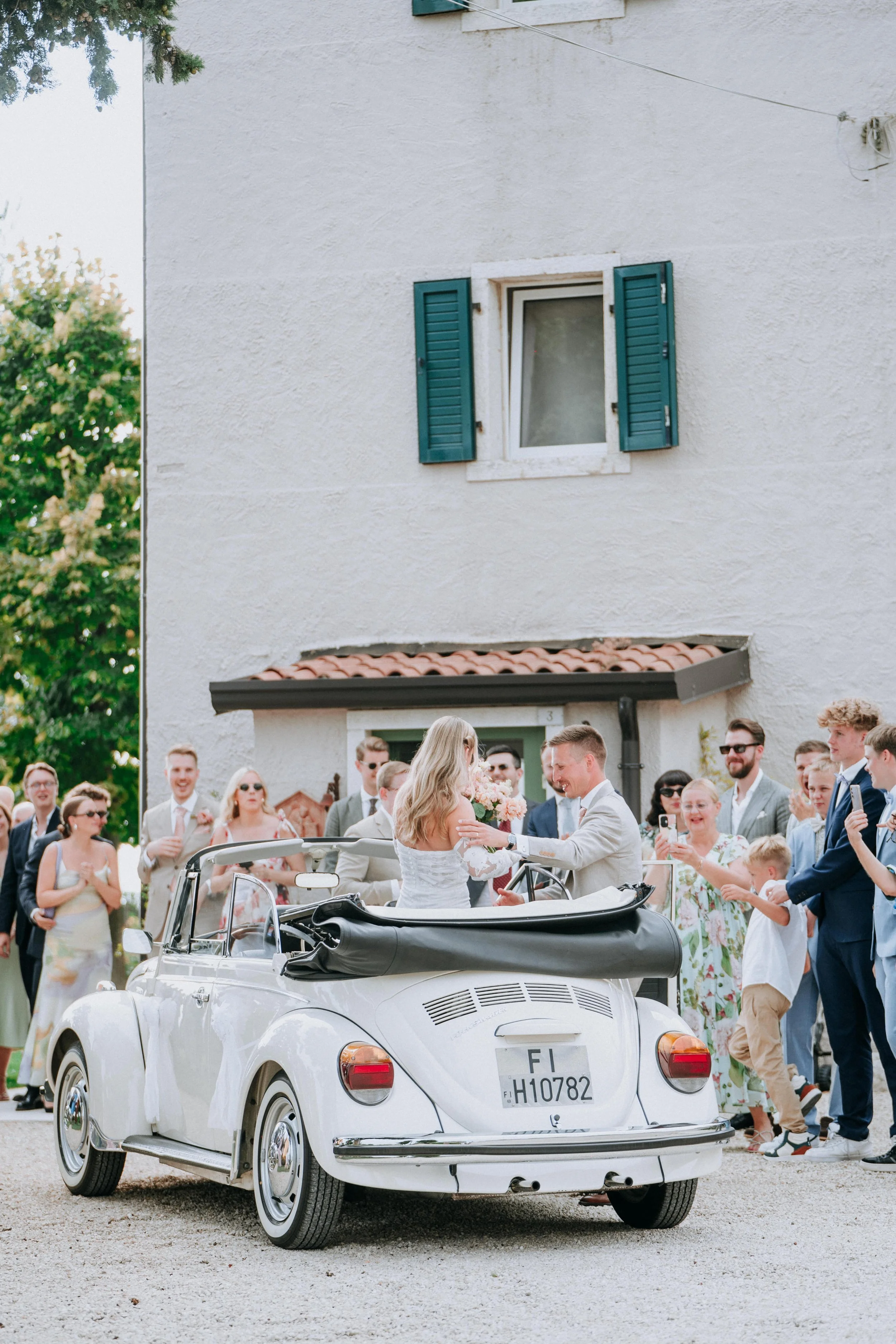 Bride and groom riding in a vintage convertible car at their wedding, surrounded by wedding guests taking photos.