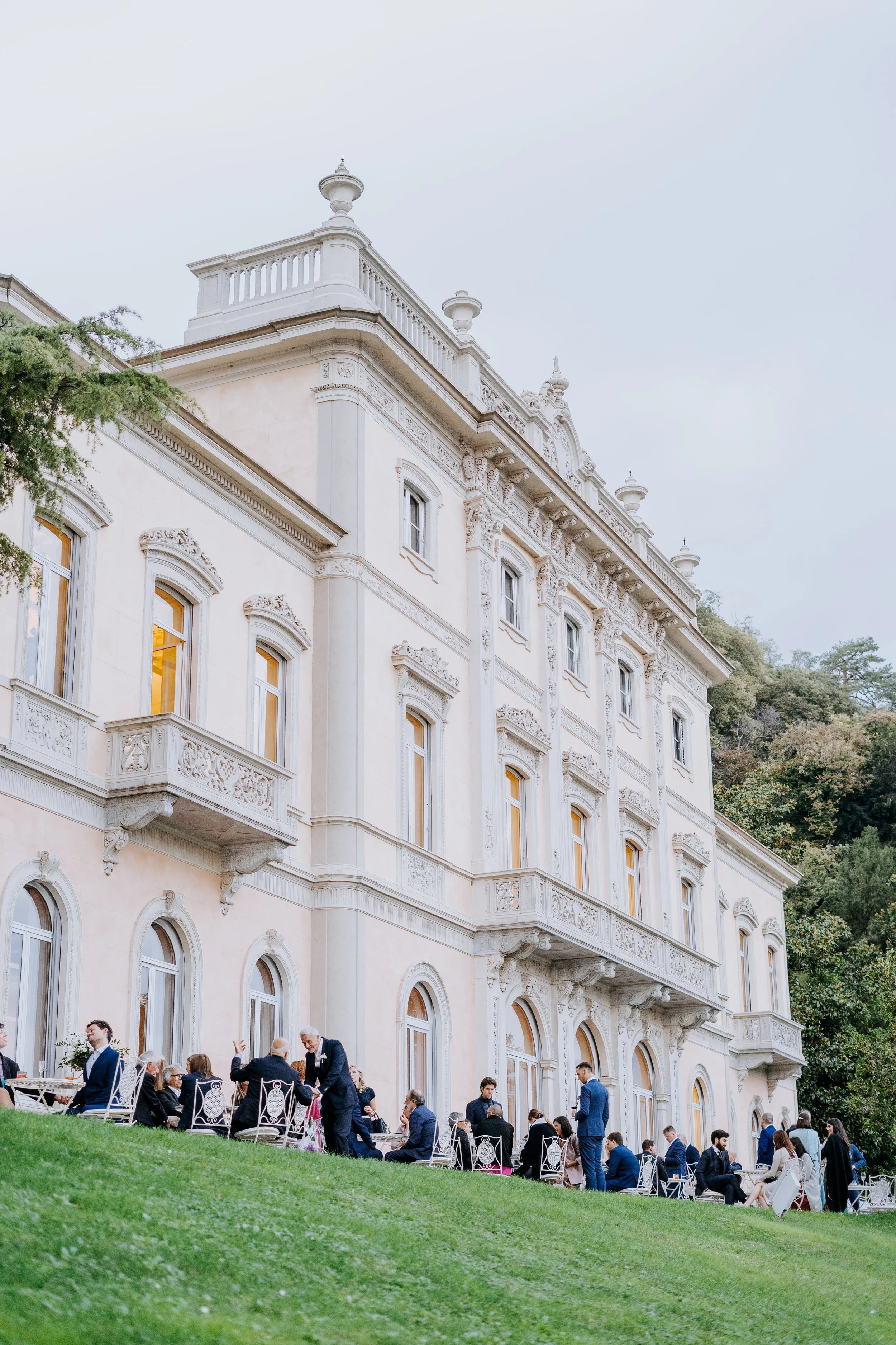 A large, ornate, white mansion with multiple floors, detailed architecture, and balconies, set against a backdrop of trees. In front, people in formal attire are gathered on a grassy lawn, sitting and standing, engaged in conversation, possibly at an