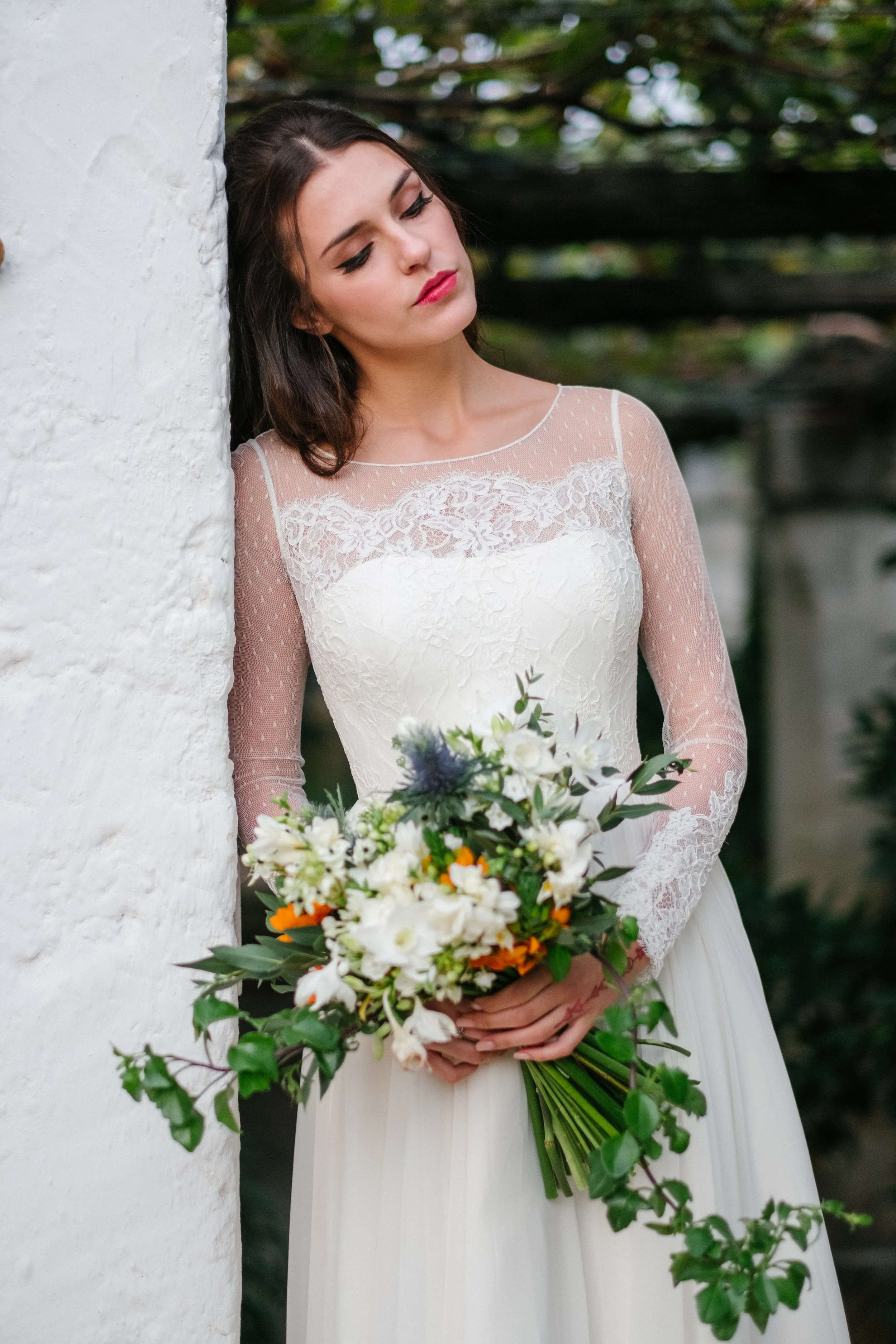 A woman in a white lace wedding dress holding a bouquet of white, orange, and purple flowers, standing against a white wall with greenery in the background.