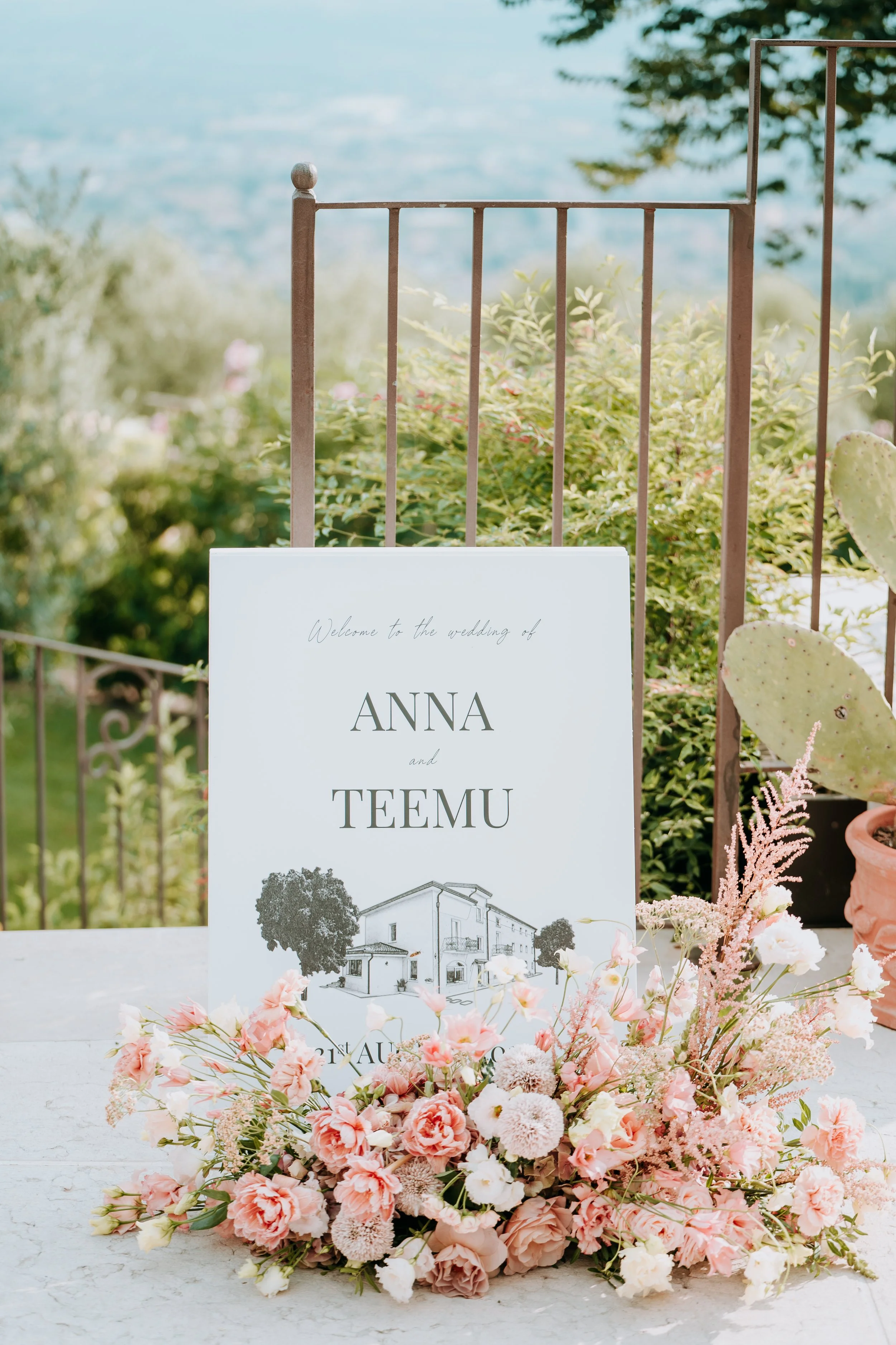 Wedding welcome sign for Anna and Teemu decorated with pink and white flowers, set outdoors on a table with greenery and cactus plants in the background.