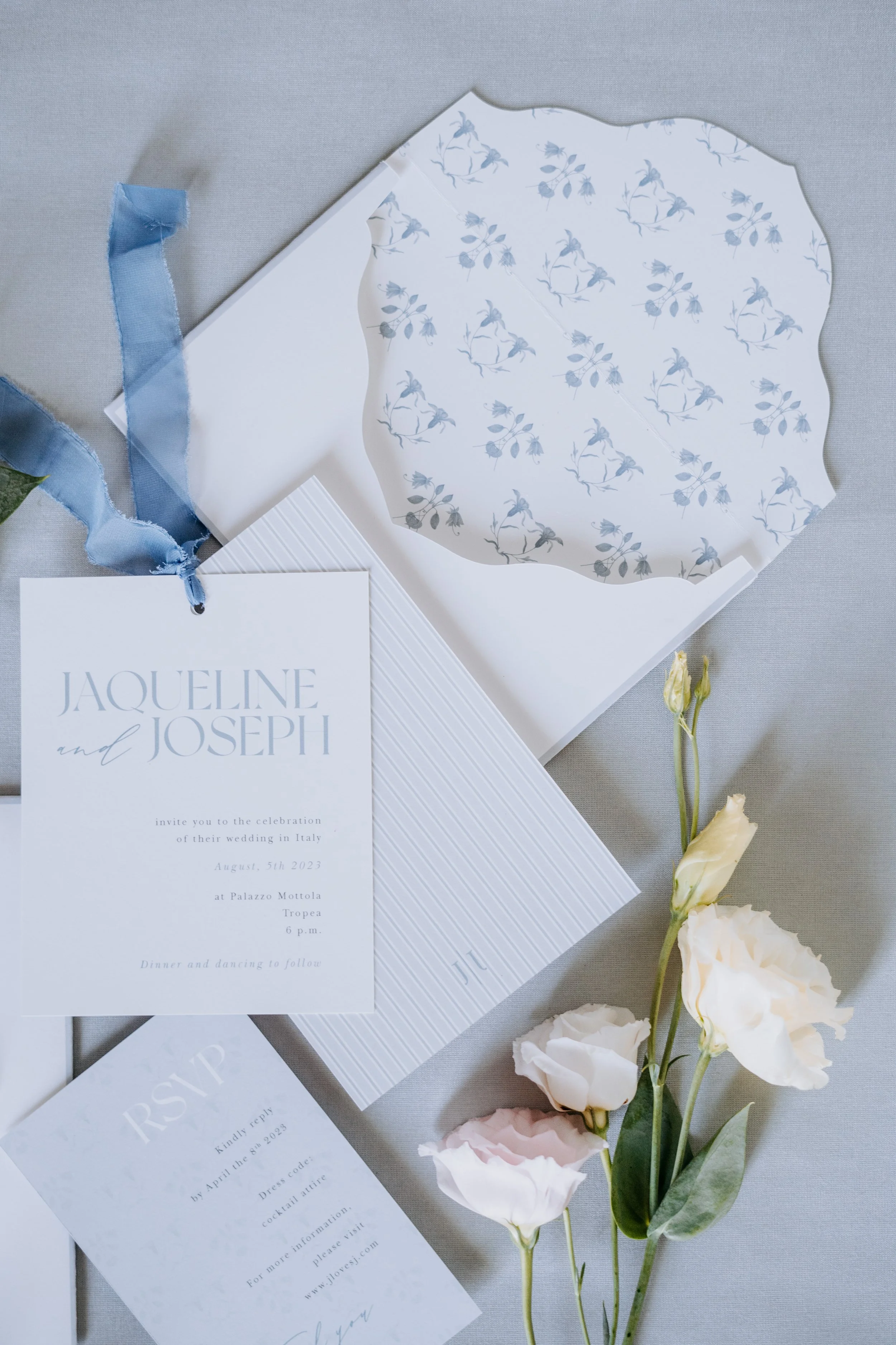 Wedding invitation suite with a floral design, RSVP card, and a blue ribbon, along with white and pale pink flowers on a gray surface.
