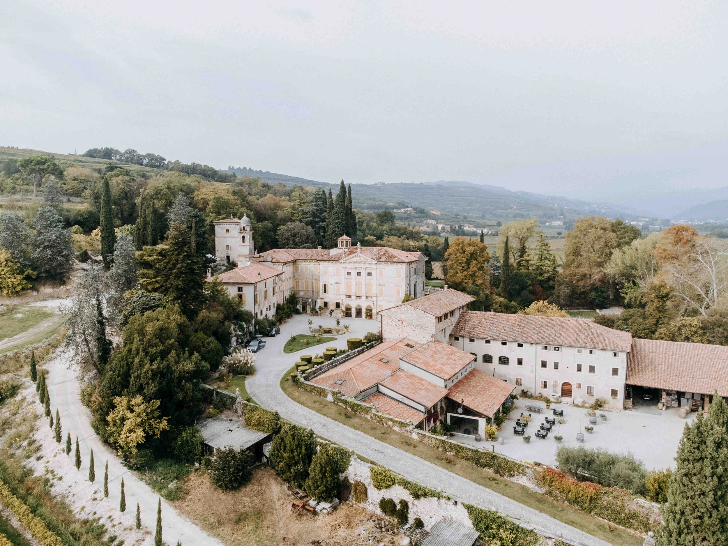 Aerial view of a historic estate with multiple stone buildings, surrounded by greenery, cypress trees, and rolling hills under a cloudy sky.