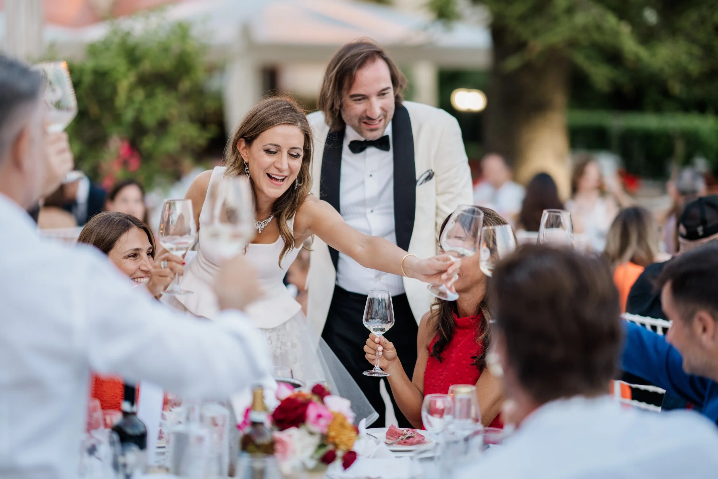 A group of people celebrating at a wedding reception, with a woman in a white dress and a man in a tuxedo serving drinks to guests at a decorated outdoor table.