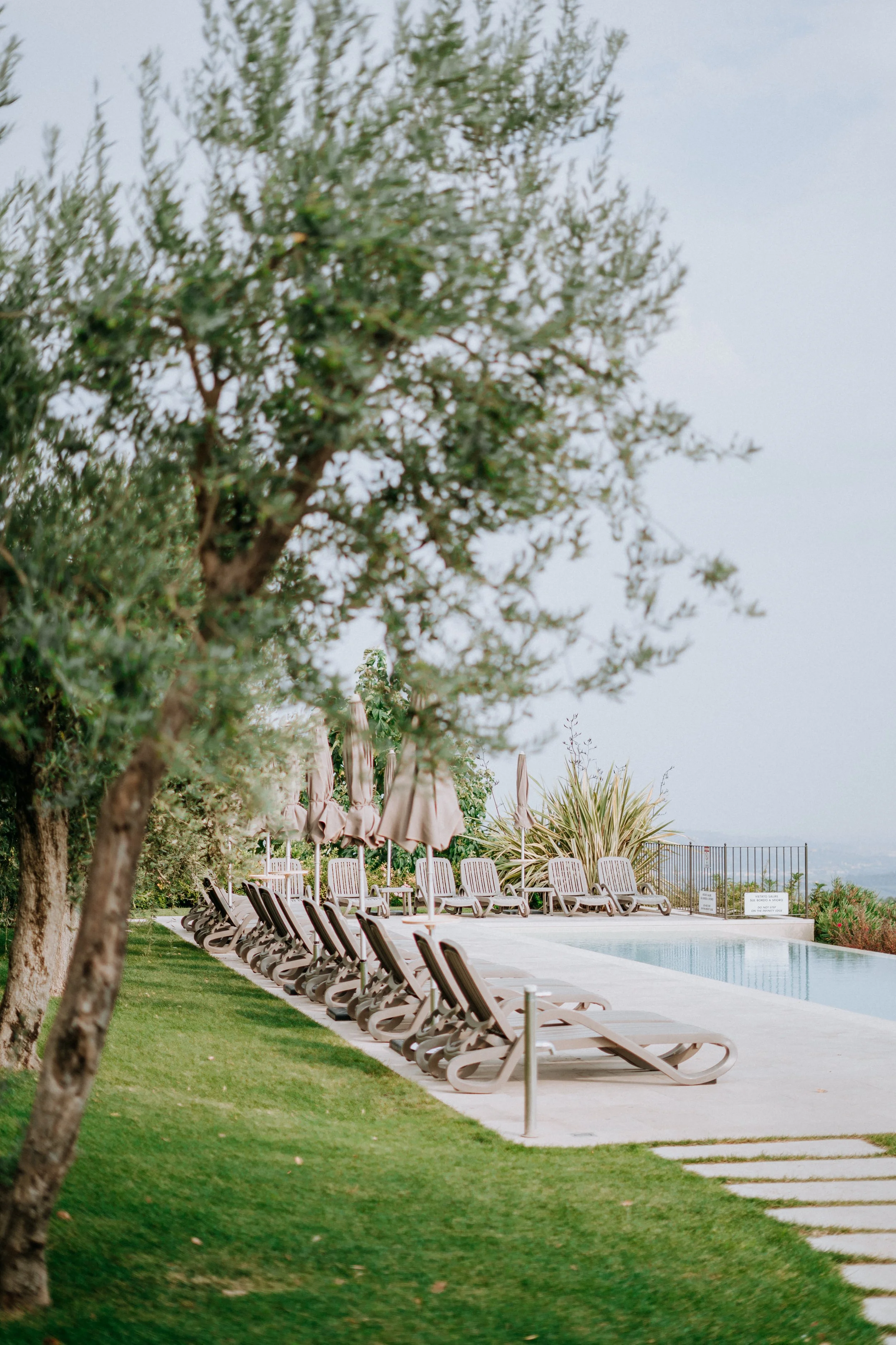 Poolside area with lounge chairs and umbrellas, surrounded by grass, trees, and decorative plants.