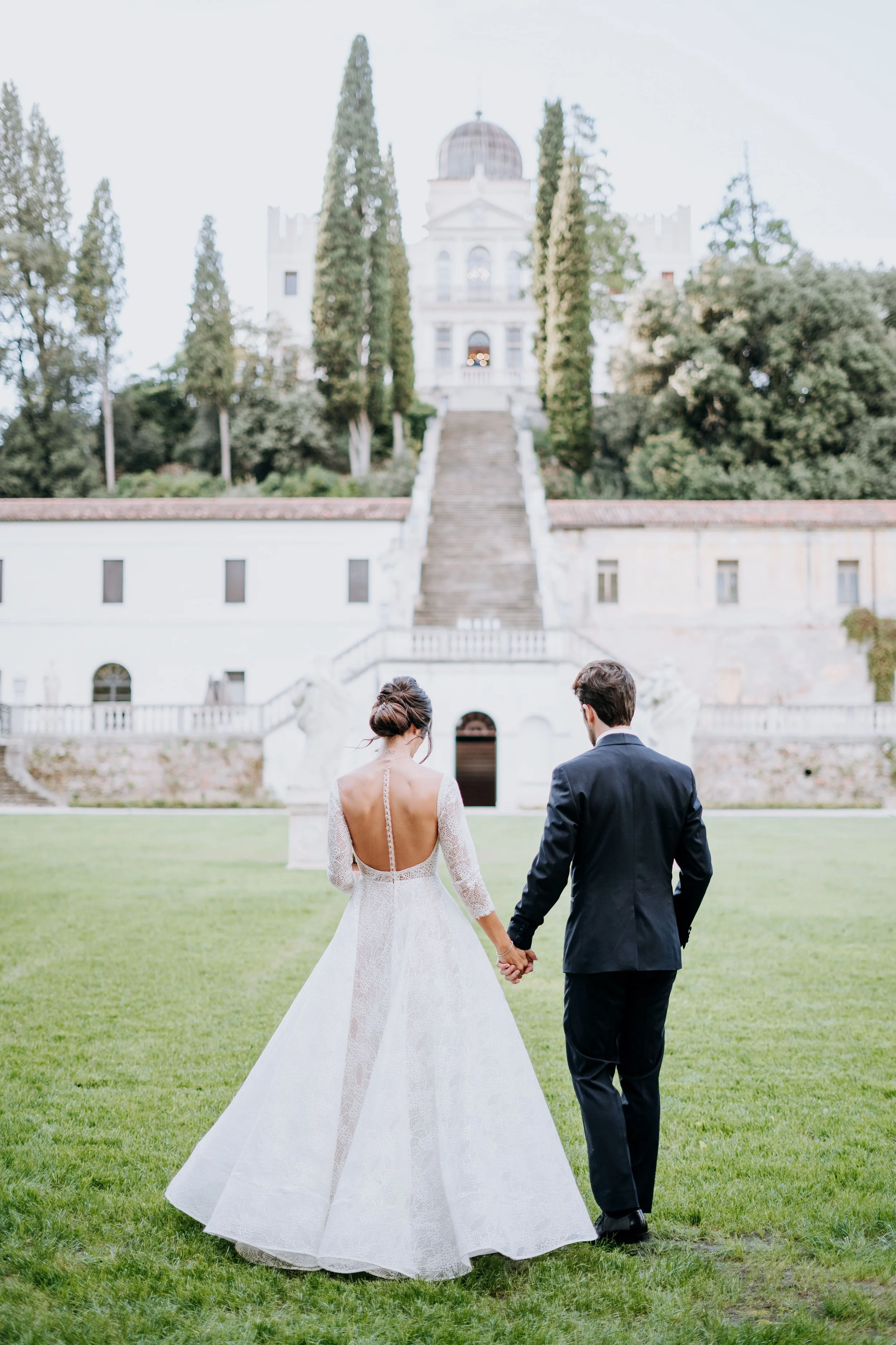 A bride and groom holding hands and walking towards a large historic building on a green lawn.