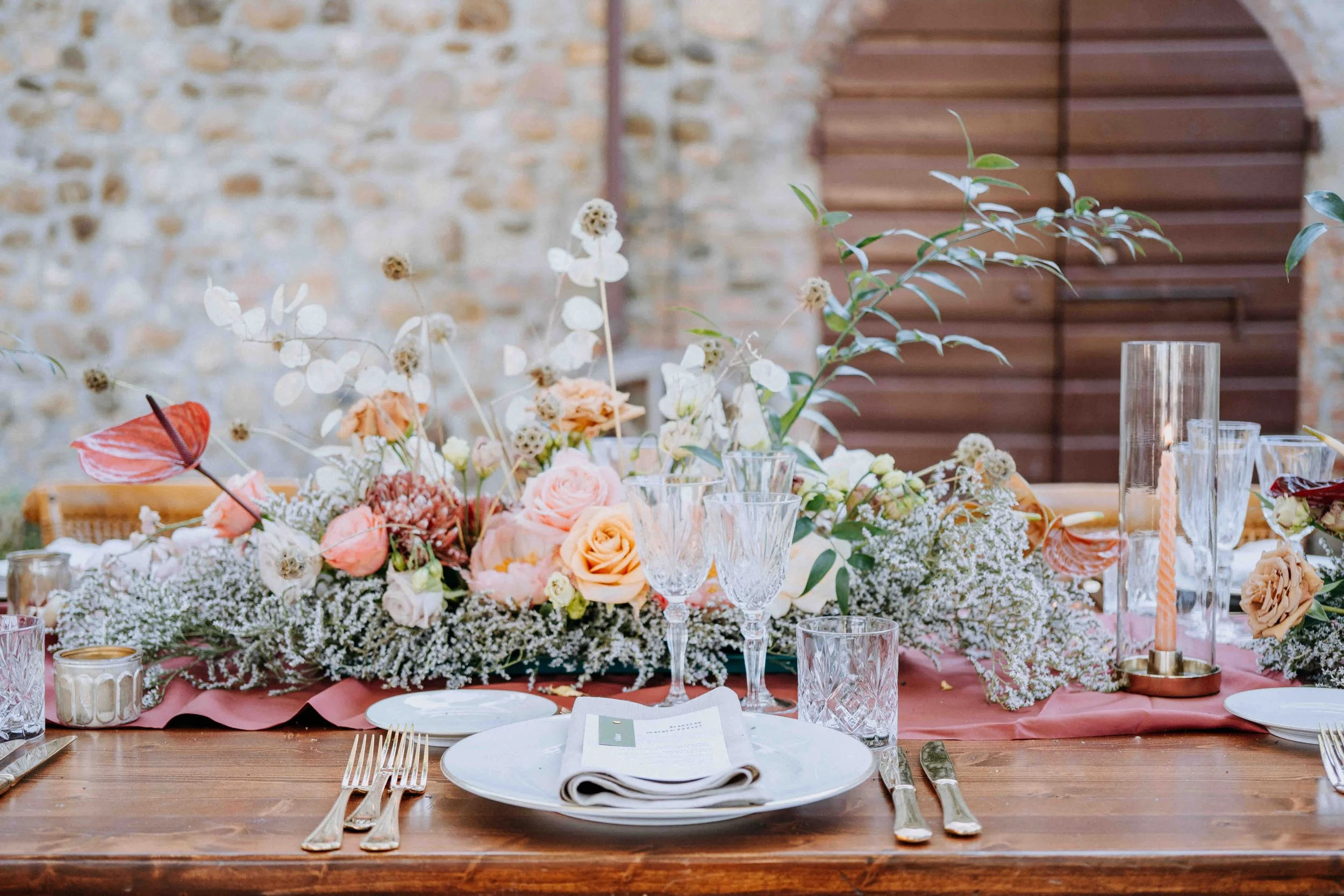 A decorated dining table with a floral centerpiece, wine glasses, candles, and place settings, set against a brick wall and wooden background at an outdoor event.