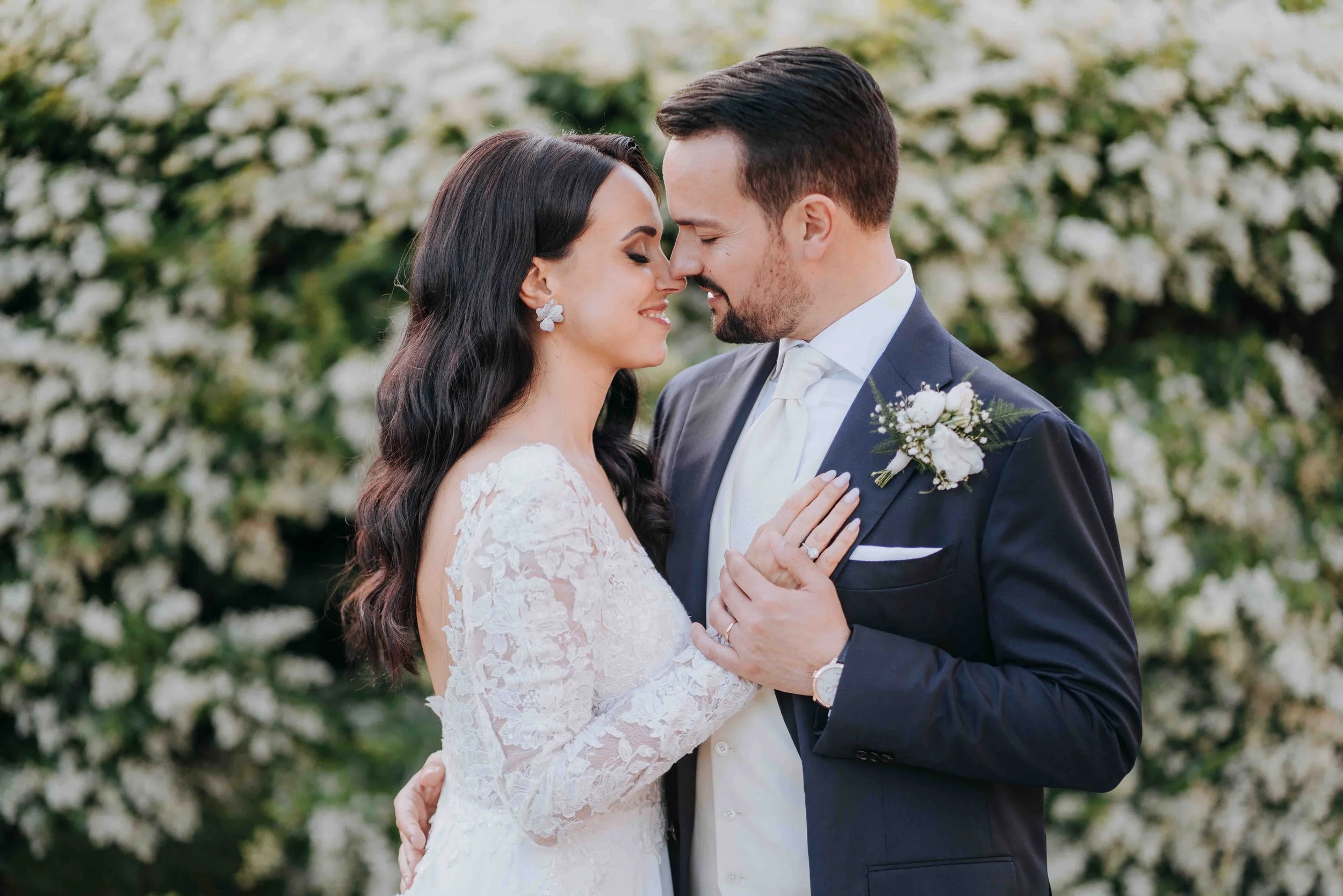 A bride and groom standing close, touching foreheads with eyes closed, outside with white flowers in the background, during their wedding.