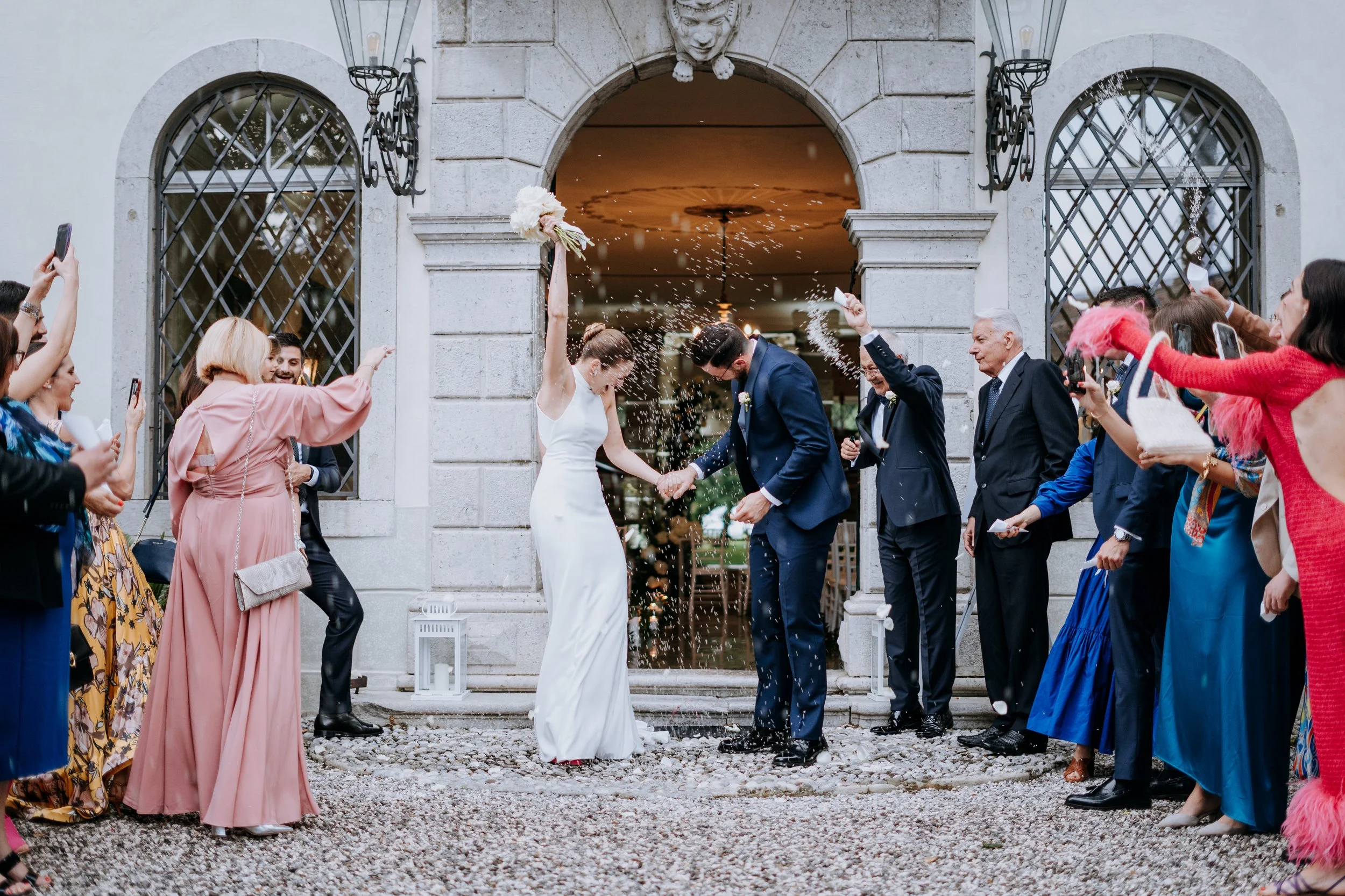 A newlywed couple celebrating outside a building with family and friends throwing confetti and taking photos.
