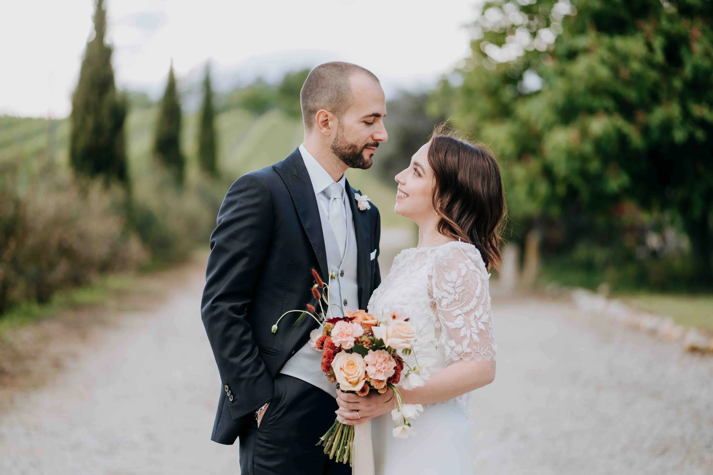 A bride and groom standing outdoors on a wedding day, gazing at each other, with the bride holding a bouquet of flowers.