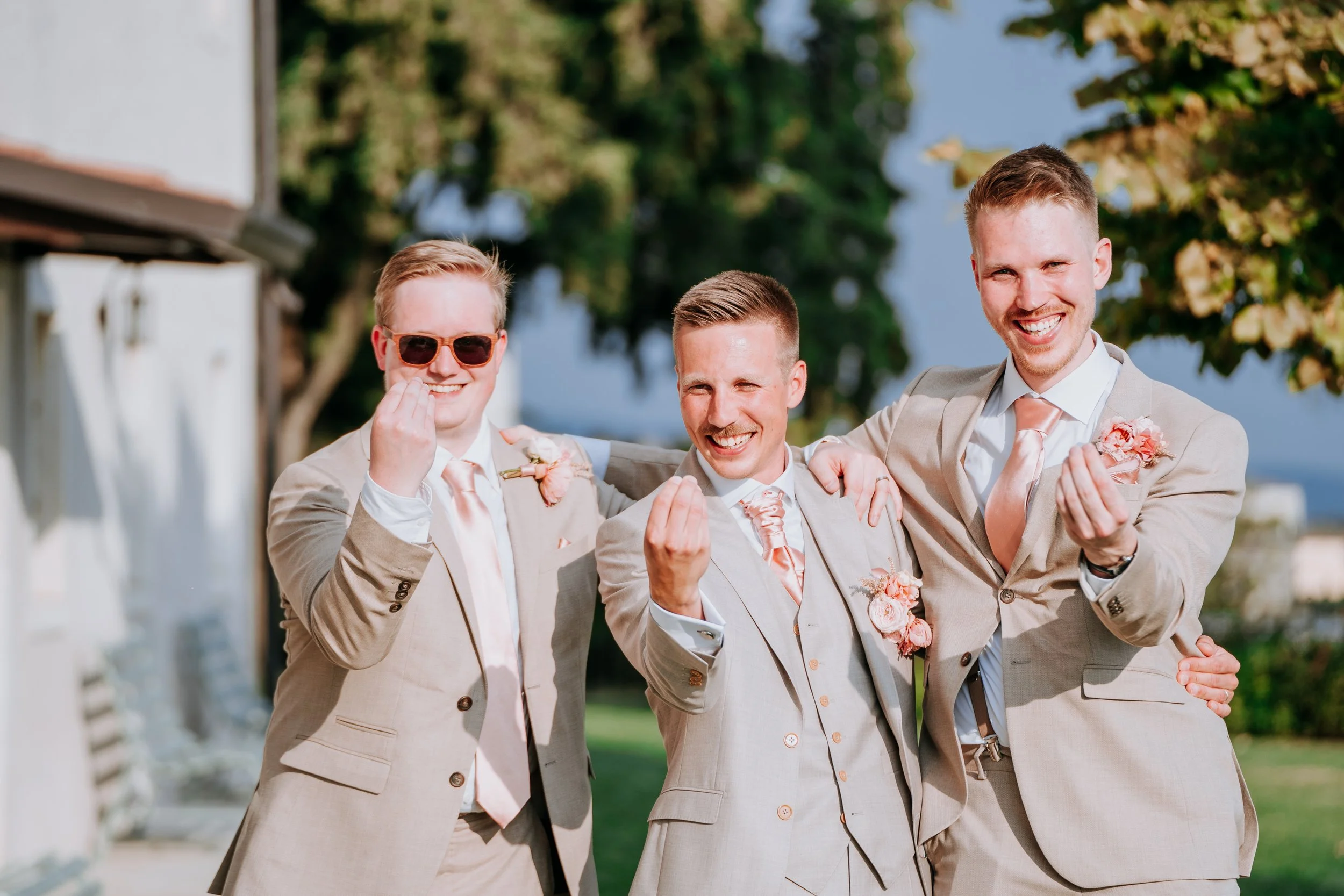 Group of three smiling men in beige suits with pink ties and boutonnieres, making finger heart gestures outdoors on a sunny day.