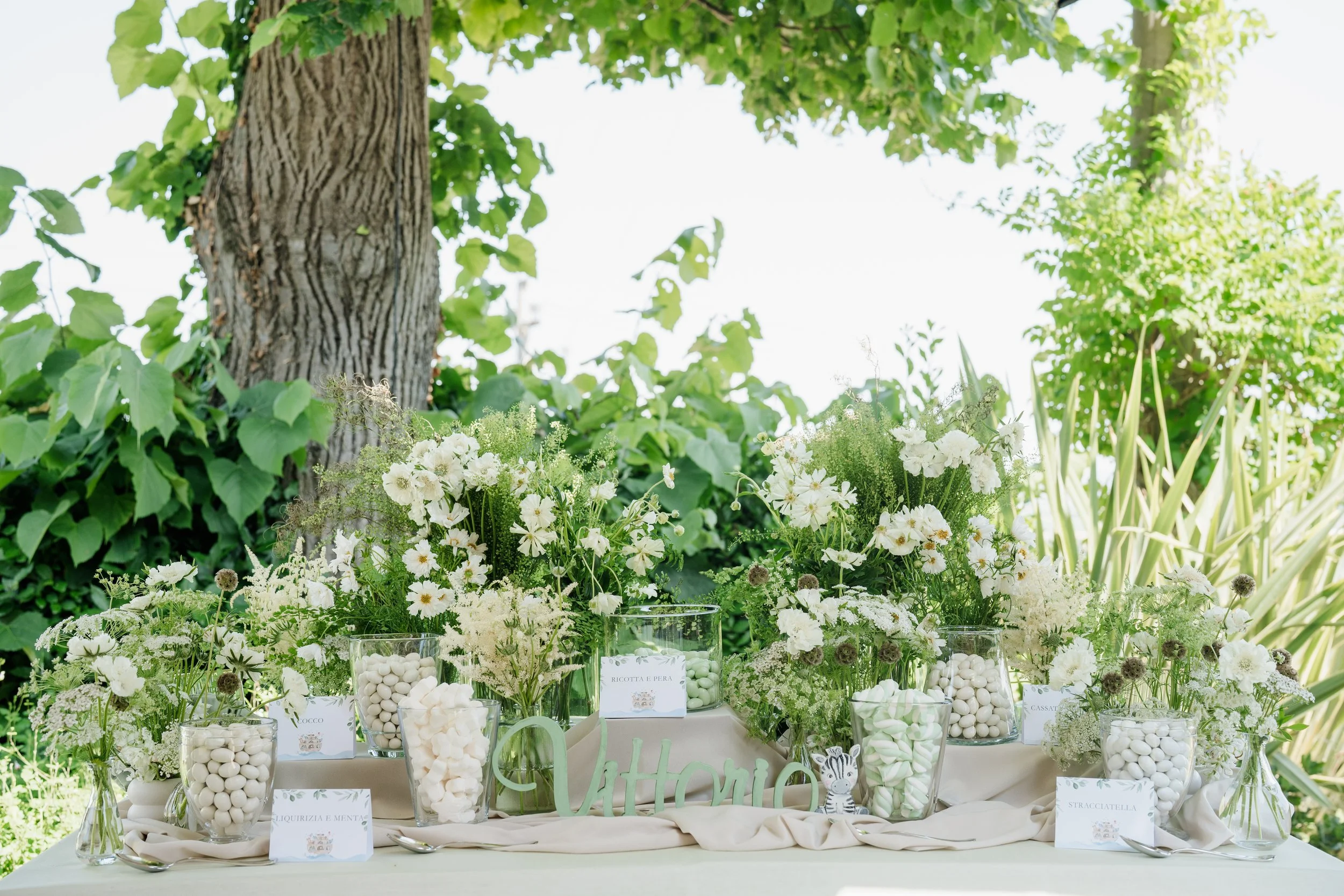 A table decorated with an arrangement of white flowers and glass jars filled with white candies, set outdoors under a large leafy tree.